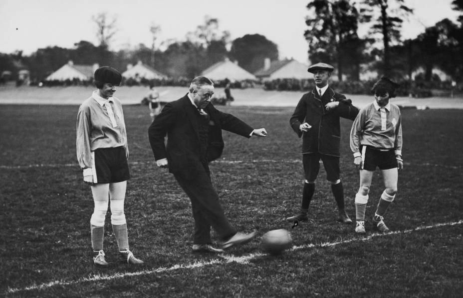 As meninas do Dick, Kerr's Ladies tinham uma agenda lotada, jogando mais de 60 partidas em um ano. Apesar disso, elas conseguiam conciliar o trabalho na fábrica. Nesta fotografia, o time estava representando a Inglaterra em um jogo contra a França, em Herne Hill, em 1925. Nela, o músico George Robey (1869 - 1954) faz um chute contra o gol.