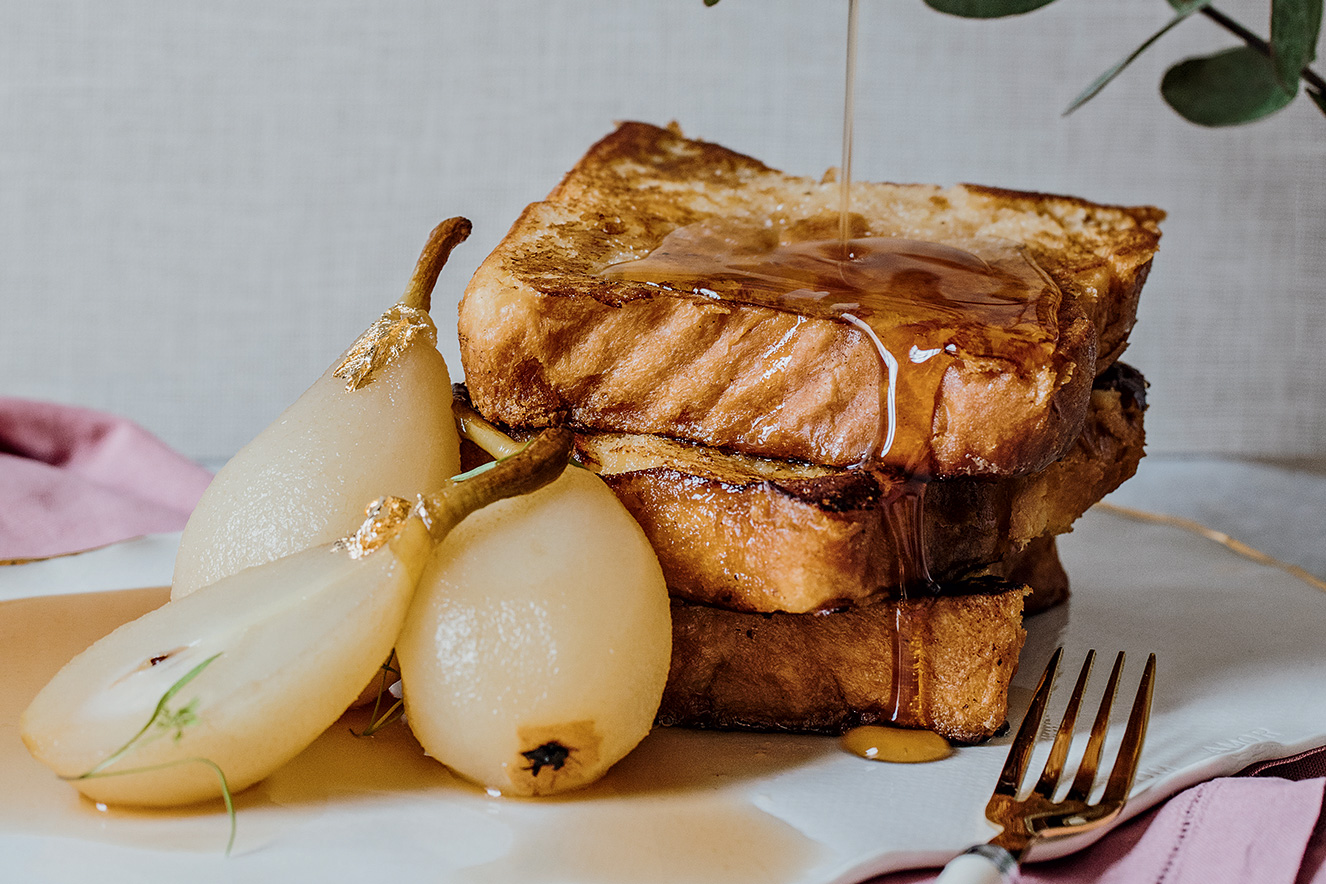 Tr&ecirc;s fatias de p&atilde;o empilhadas e cobertas por mel est&atilde;o ao lado de tr&ecirc;s peras ao vinho e sem casca. &Agrave; frente, aparece um garfo