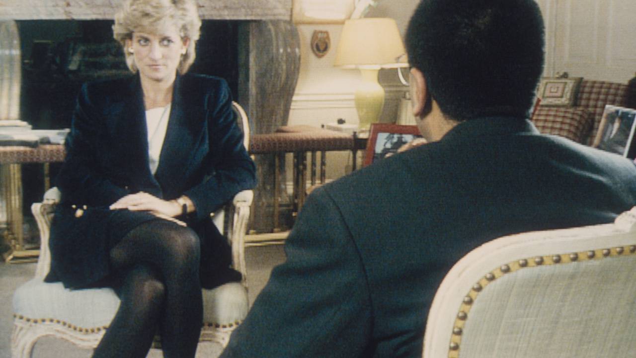 Diana, uma mulher branca com os cabelos loiros curtos está vestindo um blazer preto e uma blusa branca, meia-calças escuras. Ela olha para um homem que está de costas na foto, também de blazer escuro.