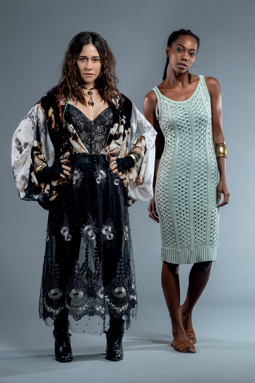 Duas mulheres posam para a c&acirc;mera. Uma, branca, com o cabelo castanho e solto, usa um vestido fluido preto florido. Ela est&aacute; com as m&atilde;os na cintura. A outra, negra, com o cabelo preso em duas tran&ccedil;as, est&aacute; com um vestido verde.