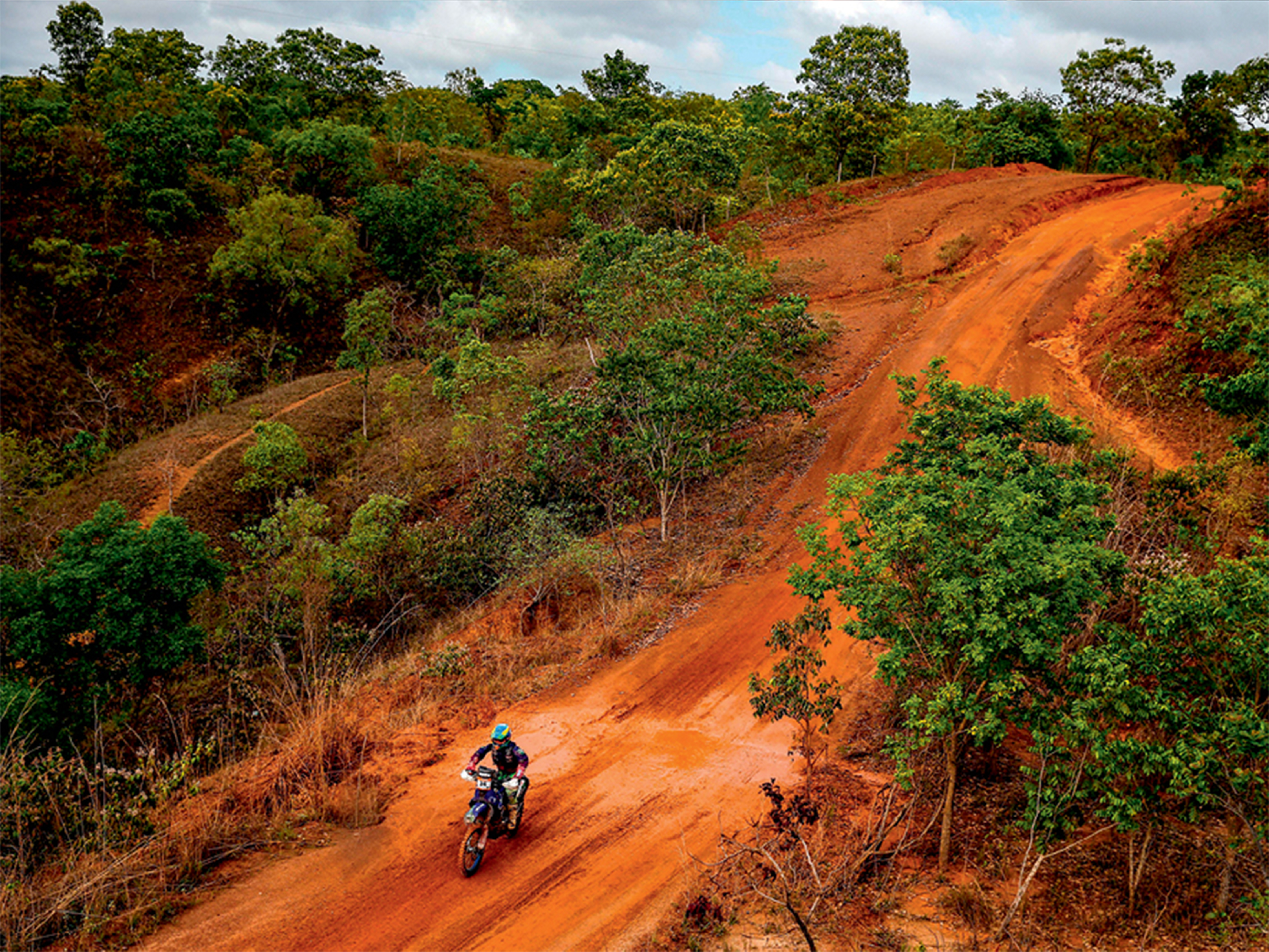 Uma faixa aberta em meio &agrave; vegeta&ccedil;&atilde;o vira pista no cen&aacute;rio de terra, com ch&atilde;o vermelho. Uma moto, pilotada por uma mulher, aparece no centro da pista