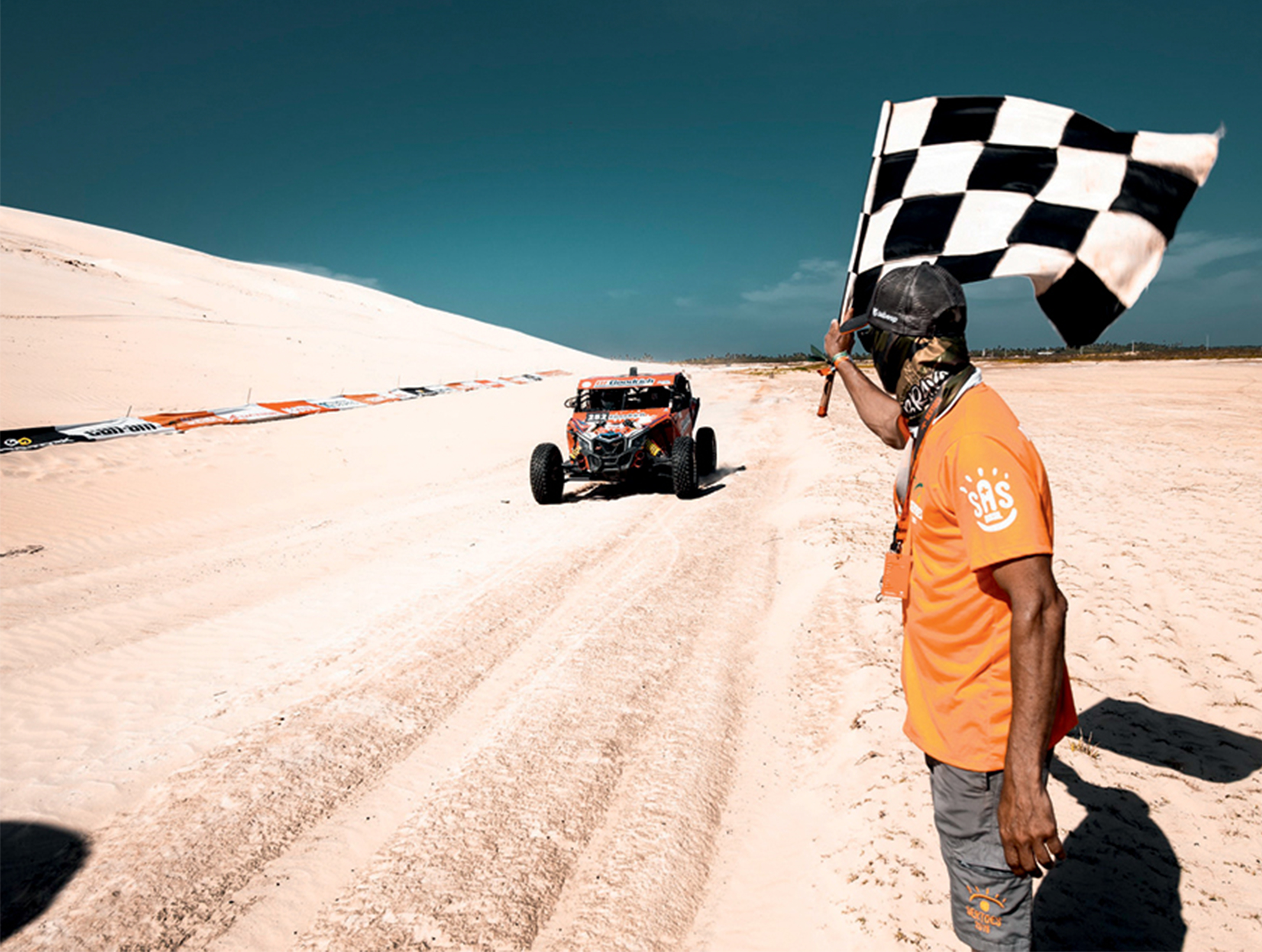 Em um terreno arenoso bem claro, um carro corre e se aproxima da bandeirada, dada por um homem de camiseta laranja