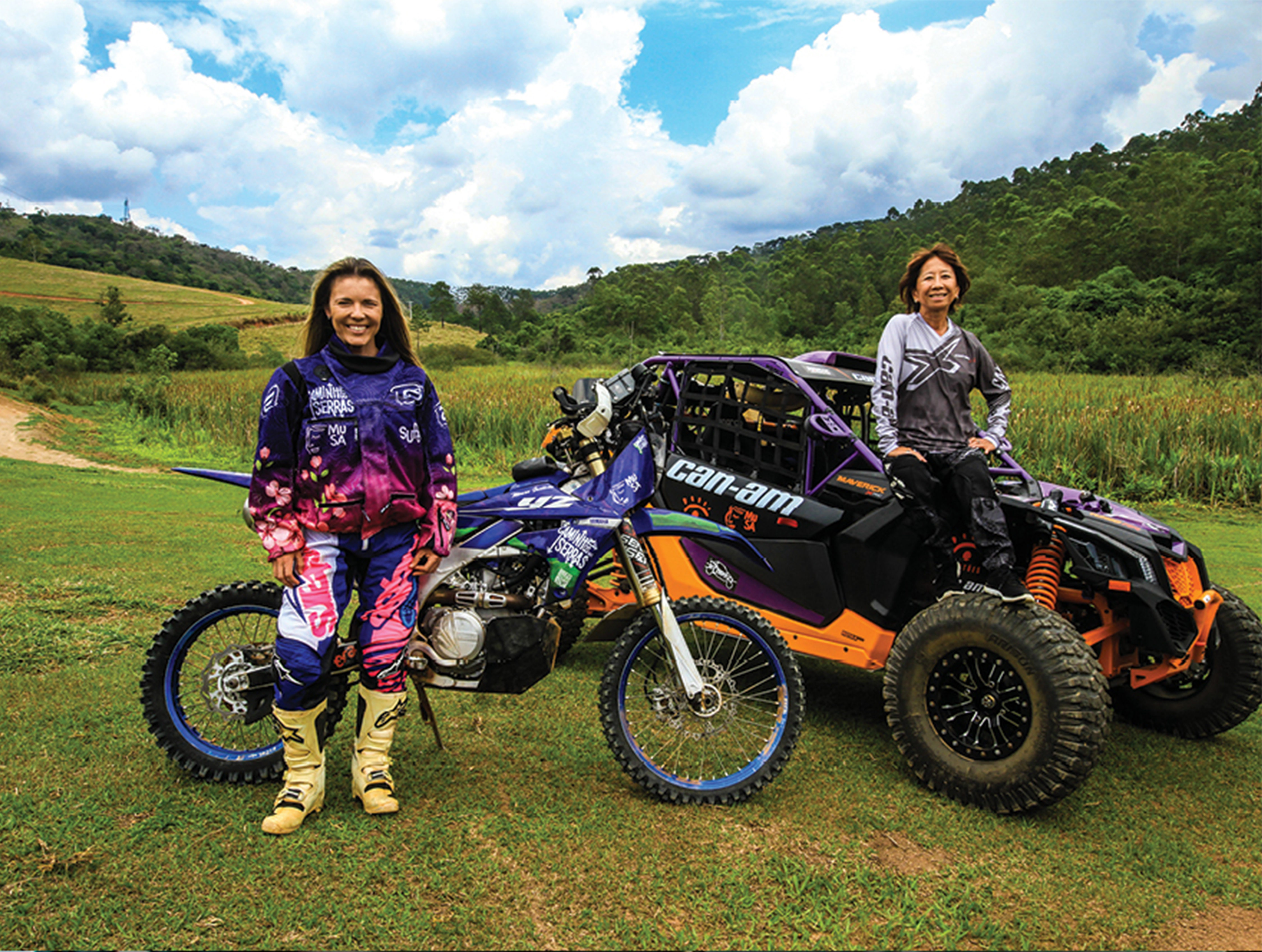 Em um campo verde, duas mulheres vestidas com uniformes est&atilde;o paradas junto aos seus ve&iacute;culos. Uma delas, loira, tem uma moto, usa uniforme rosa e roxo e botas amarelas. A outra, de cabelos castanhos curtos, usa uniforme cinza e preto e est&aacute; sentada sobre um UTV