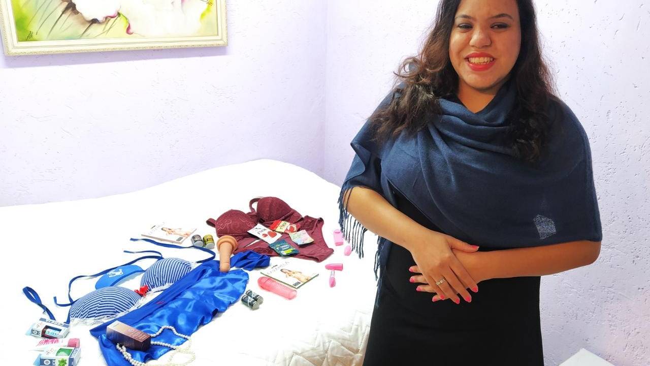 Thais Coutinho aparece de pé na frente e atrás dela estão os produtos sexuais vendidos em sua loja Blind Sexy