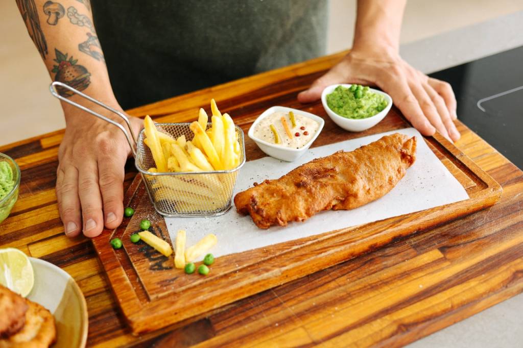 Receita de fish and chips (peixe e fritas) Mãos masculinas servindo fish and chips em tábua de madeira.