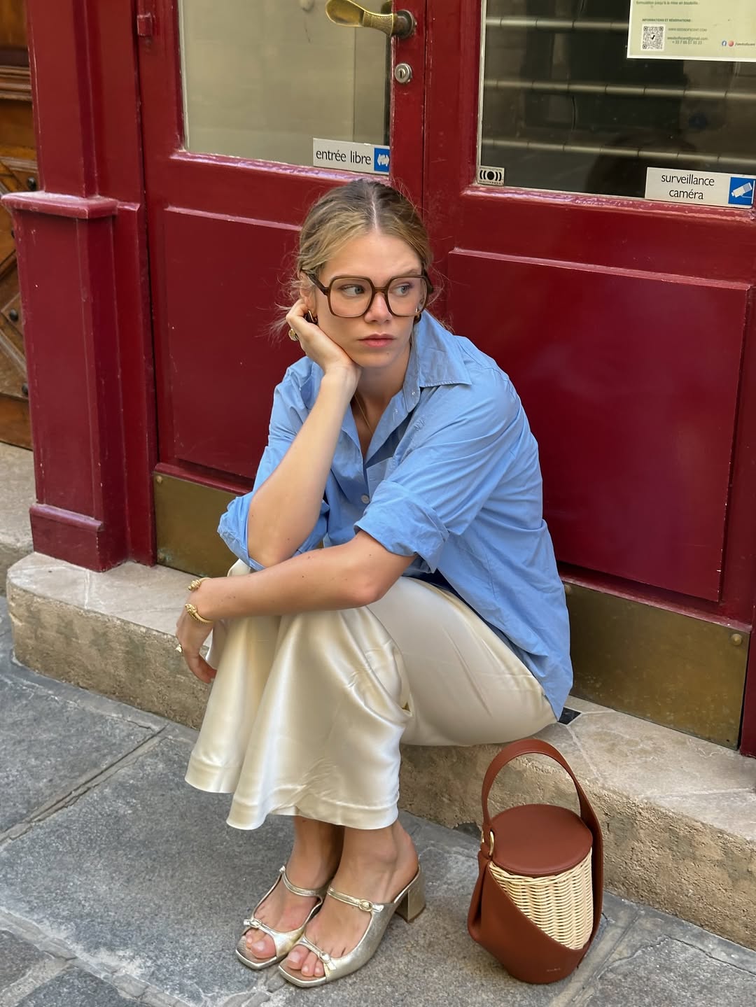 Foto de uma mulher loira usando um look composto por camisa azul, saia e salto prata, al&eacute;m de uma bolsa marrom