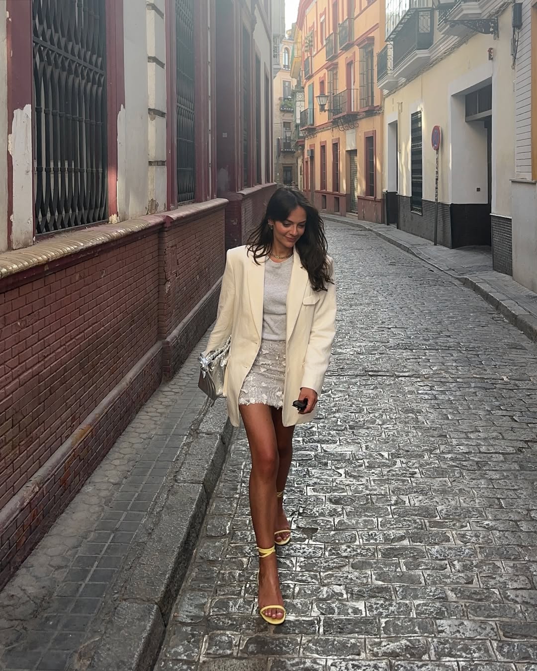 Foto de uma mulher morena usando um look composto por vestido branco, blazer branco, salto dourado e bolsa