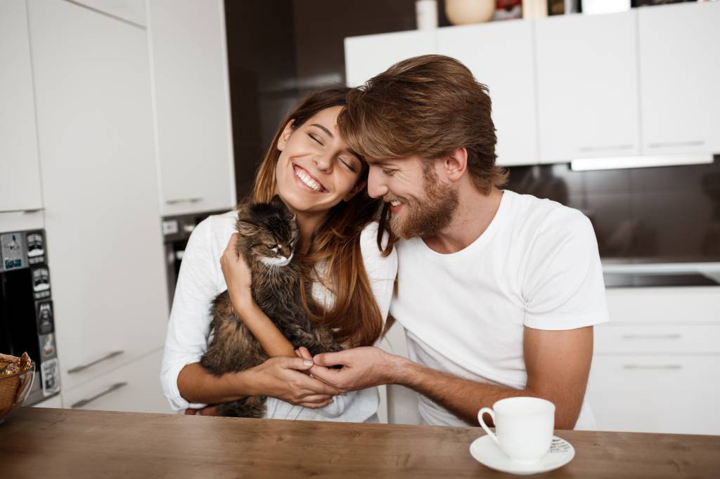 Happy young beautiful couple sitting at kitchen holding cat smiling. Homem e mulher sentados sorrindo em uma cozinha, a mulher está segurando um gato