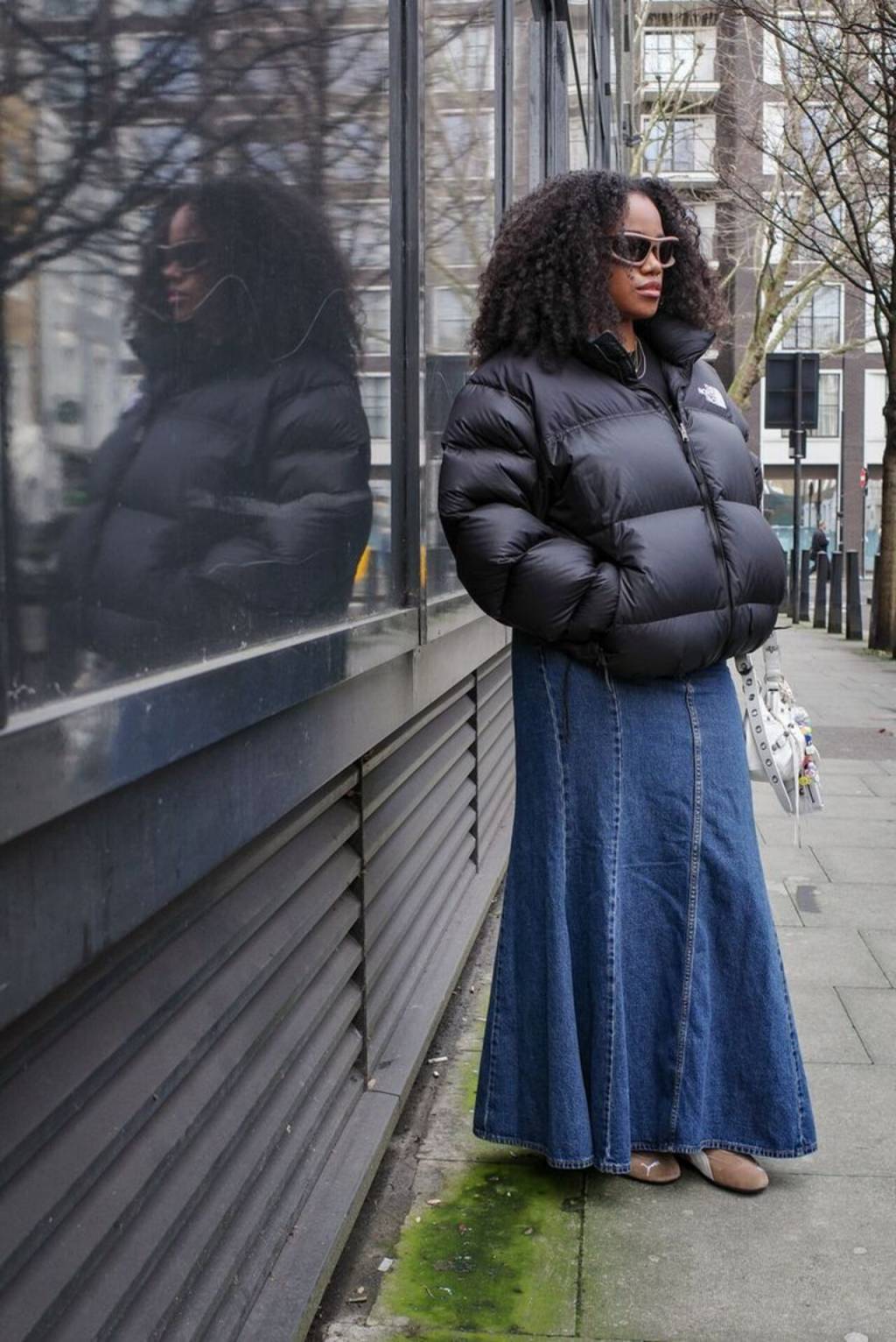 jaqueta-puffer (8) Imagem de mulher negra pousando com jaqueta puffer preta, saia longa jeans e tênis.