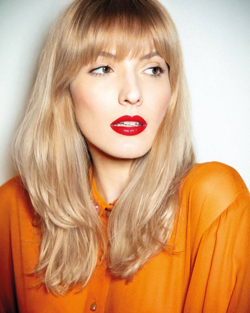 tons-cabelo-inverno Imagem de mulher branca loira com franja, batom vermelho e camiseta laranja.