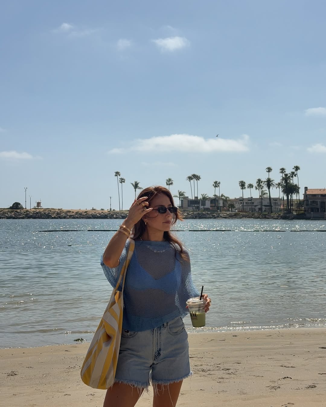 Mulher na praia com poncho azul transparente, biqu&iacute;ni azul, short jeans e bolsa listrada amarela