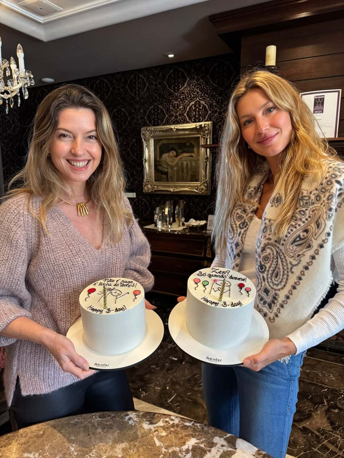 Imagem de Gisele B&uuml;ndchen segurando bolo de anivers&aacute;rio ao lado de outra mulher.