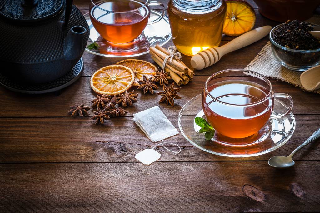 cha-de-cravo-com-laranja Xícara de chá cheia, cercada por uma colher de chá, bule, sachê de chá e alguns ingredientes, como folhas secas de chá preto, anis, laranja desidratada, paus de canela e um pote de mel. A cor predominante é o laranja.