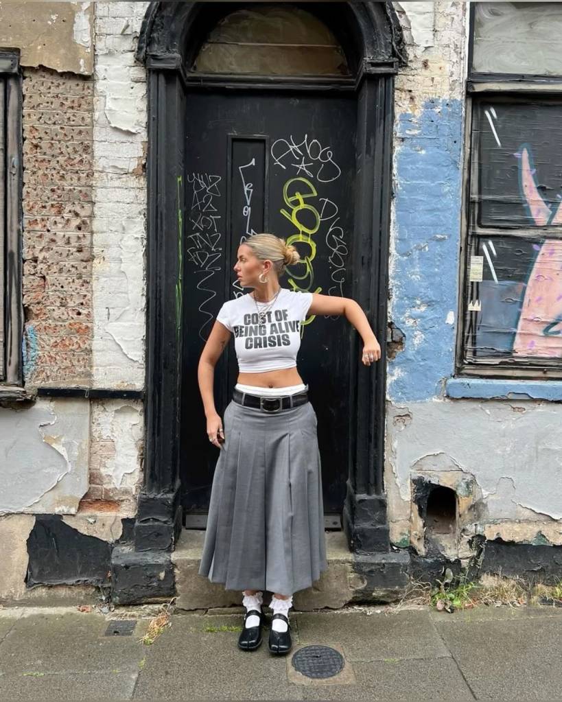 tubi-estilo-colegial Pessoa com camiseta cropped estampada, saia cinza pregueada, meia branca com babado e sapato tabi branco. Visual jovem e lúdico.