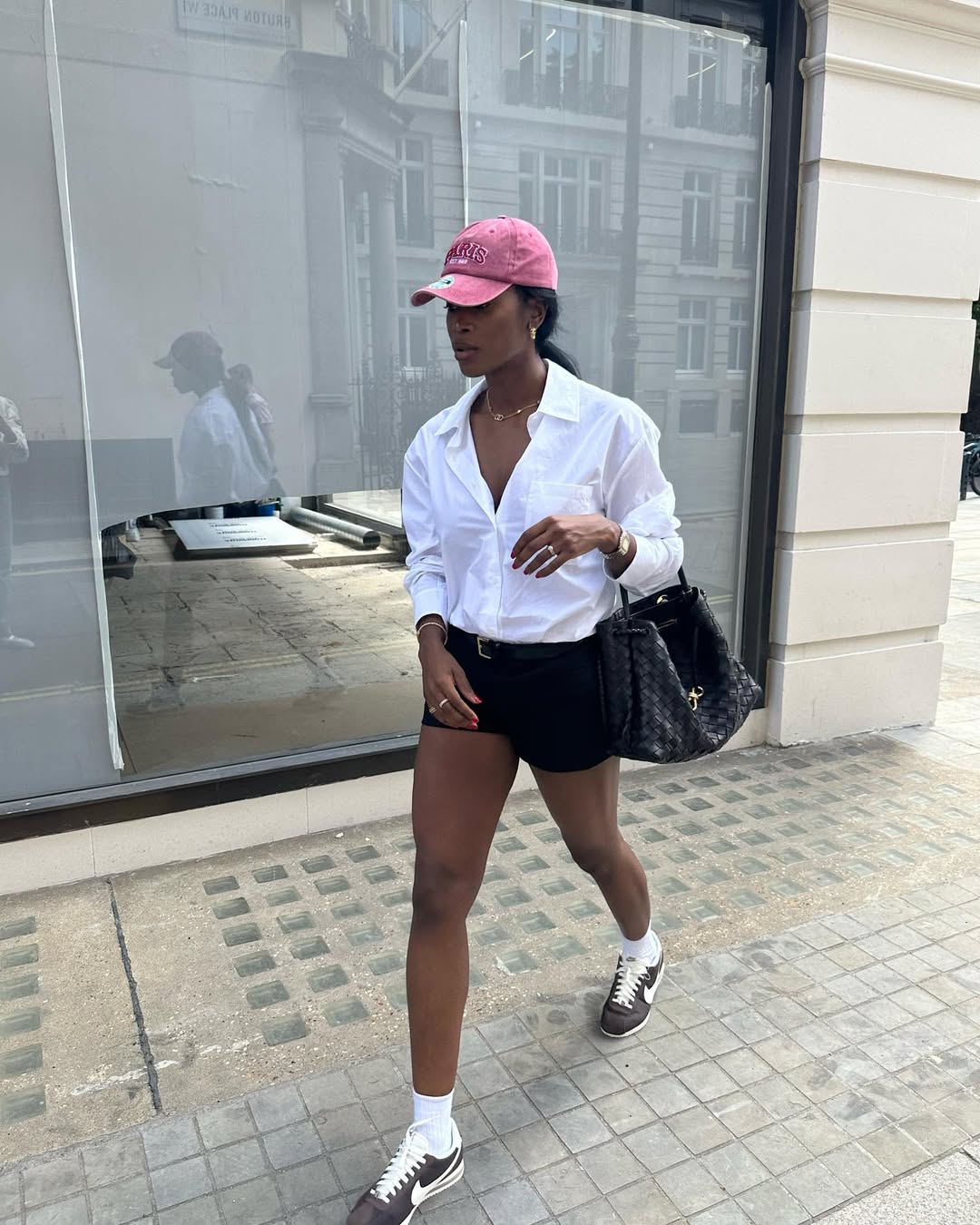 Foto de uma mulher negra usando um look composto por camisa branca, short preto, bon&eacute; rosa, bolsa preta e t&ecirc;nis marrom