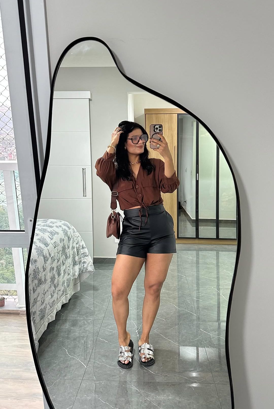 Foto de uma mulher morena usando um look composto por camisa marrom, short de couro preto, bolsa marrom e t&ecirc;nis