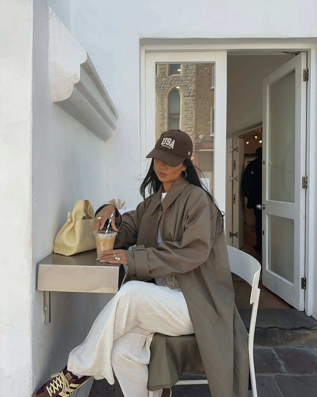 Foto de uma mulher usando um look composto por trench coat, bon&eacute;, cal&ccedil;a branca e t&ecirc;nis marrom