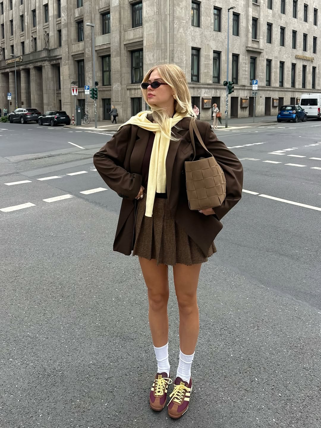Foto de uma mulher usando um look composto por blazer marrom, saia plissada marrom, t&ecirc;nis marrom e tric&ocirc; amarelo