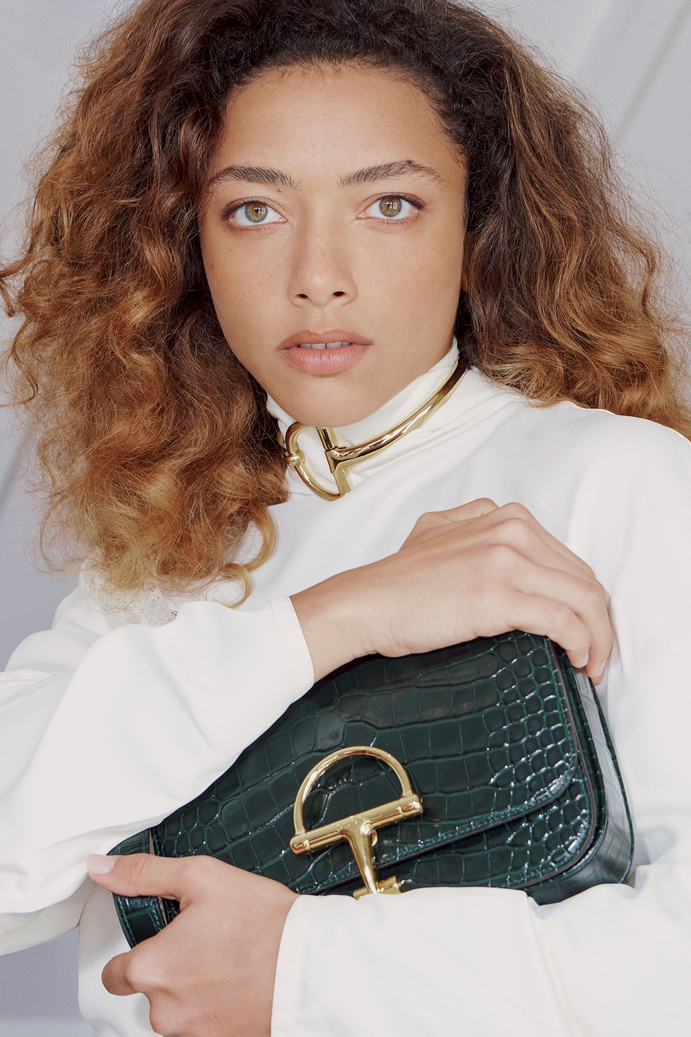 Foto de Alice Carvalho usando um look composto por blusa branca de gola alta, bolsa preta de couro e colar dourado