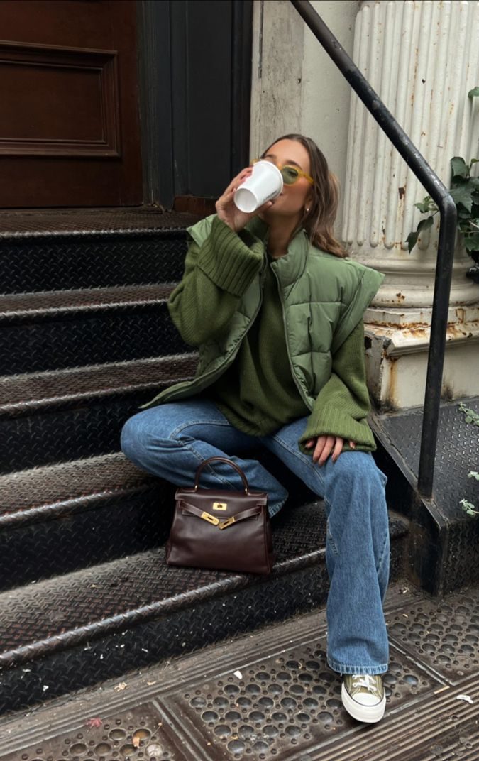 Foto de uma mulher sentada usando um look composto por blusa verde, jaqueta verde, cal&ccedil;a jeans, bolsa de couro marrom e t&ecirc;nis verde pistache