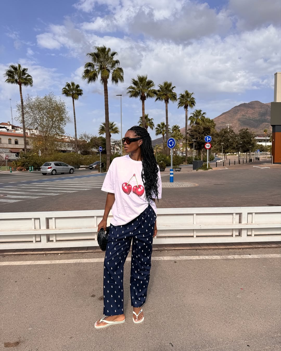 Foto de uma mulher negra usando um look composto por camiseta branca, cal&ccedil;a estampada azul, &oacute;culos de sol e bolsa preta