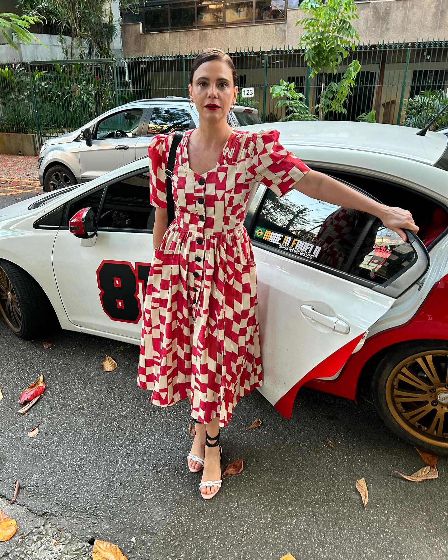 Foto de Malu Galli usando um look composto por vestido com estampas vermelhas e brancas, al&eacute;m de um salto e uma bolsa preta