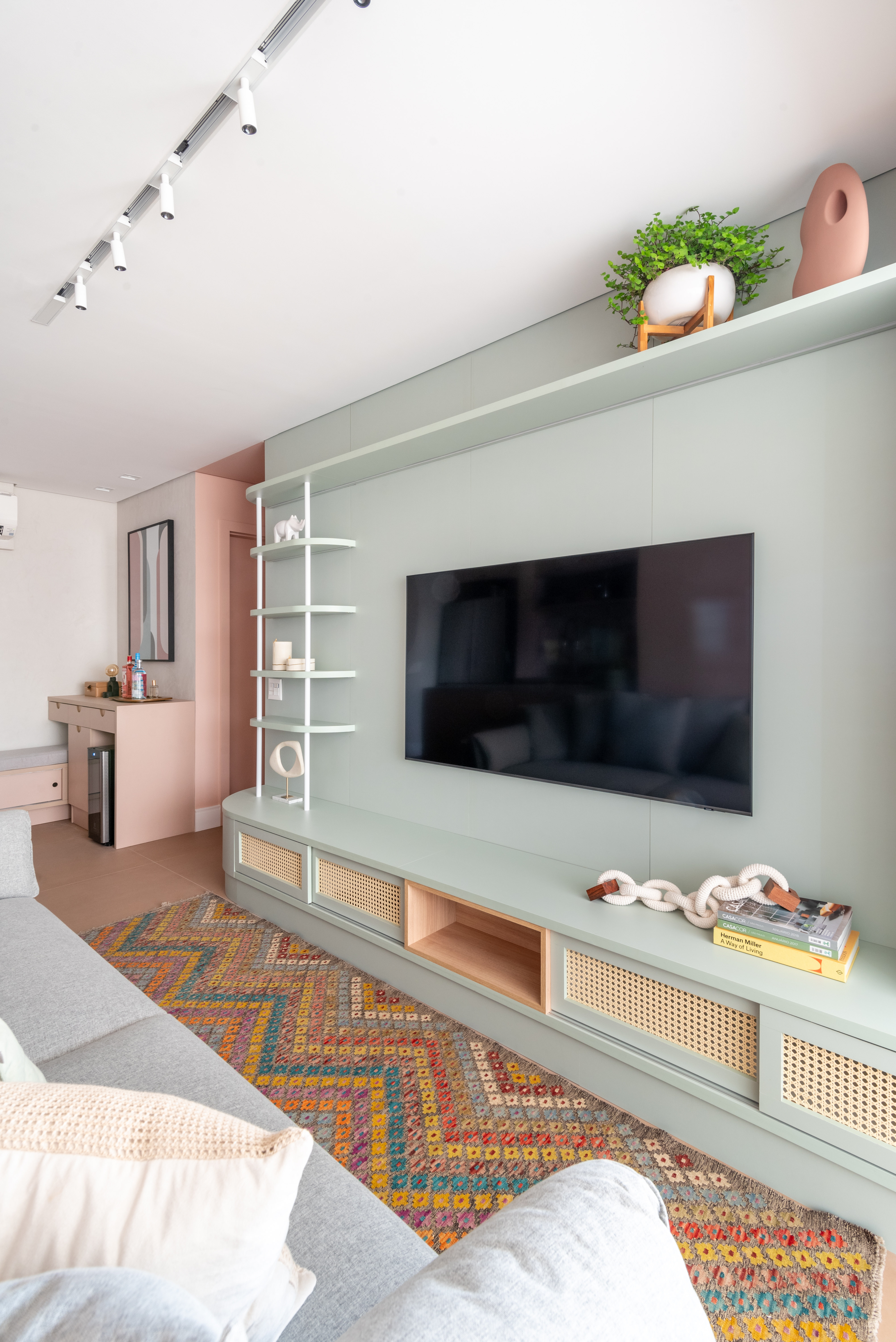 Painel de TV verde-claro com rack integrado que combina portas de palhinha, nichos de madeira e prateleiras brancas arredondadas. Sobre o m&oacute;vel aparecem livros, objetos decorativos e uma corrente escultural. Acima, vasos em cer&acirc;mica e plantas complementam a composi&ccedil;&atilde;o em uma sala iluminada e com tapete colorido geom&eacute;trico