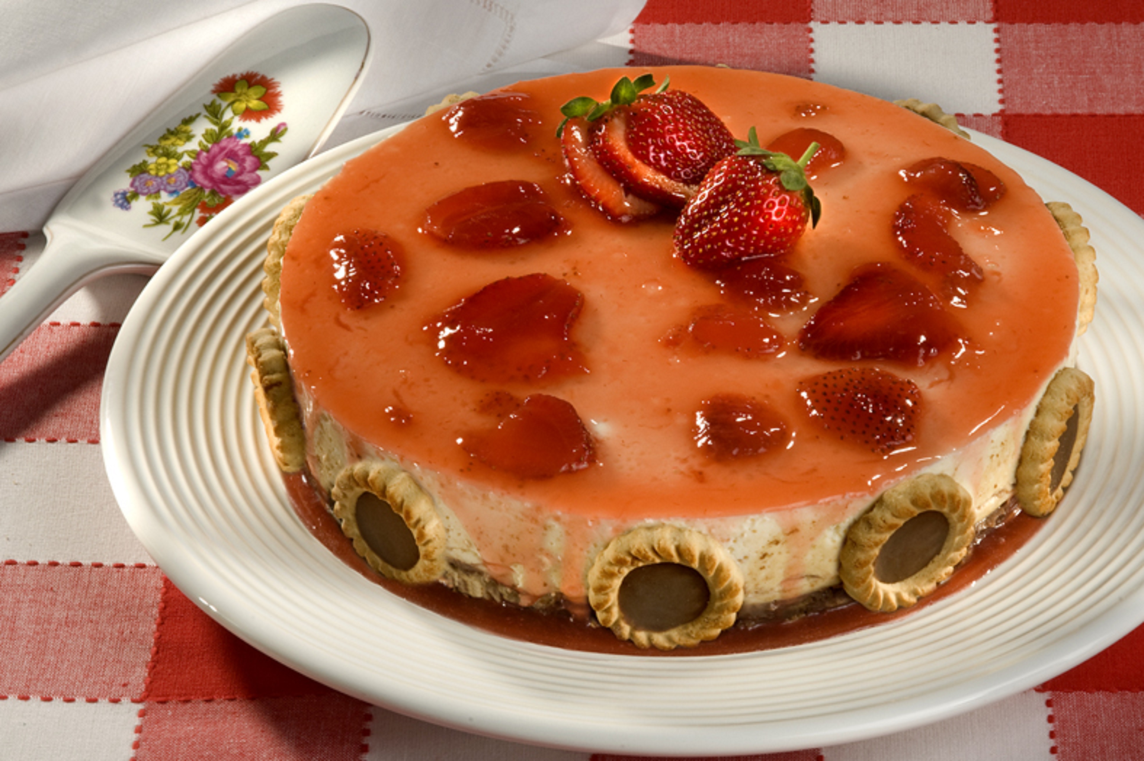 Torta gelada com camadas de creme claro e cobertura de geleia de morango, decorada com morangos frescos no topo e biscoitos recheados nas laterais. A superf&iacute;cie brilhante evidencia a textura da calda