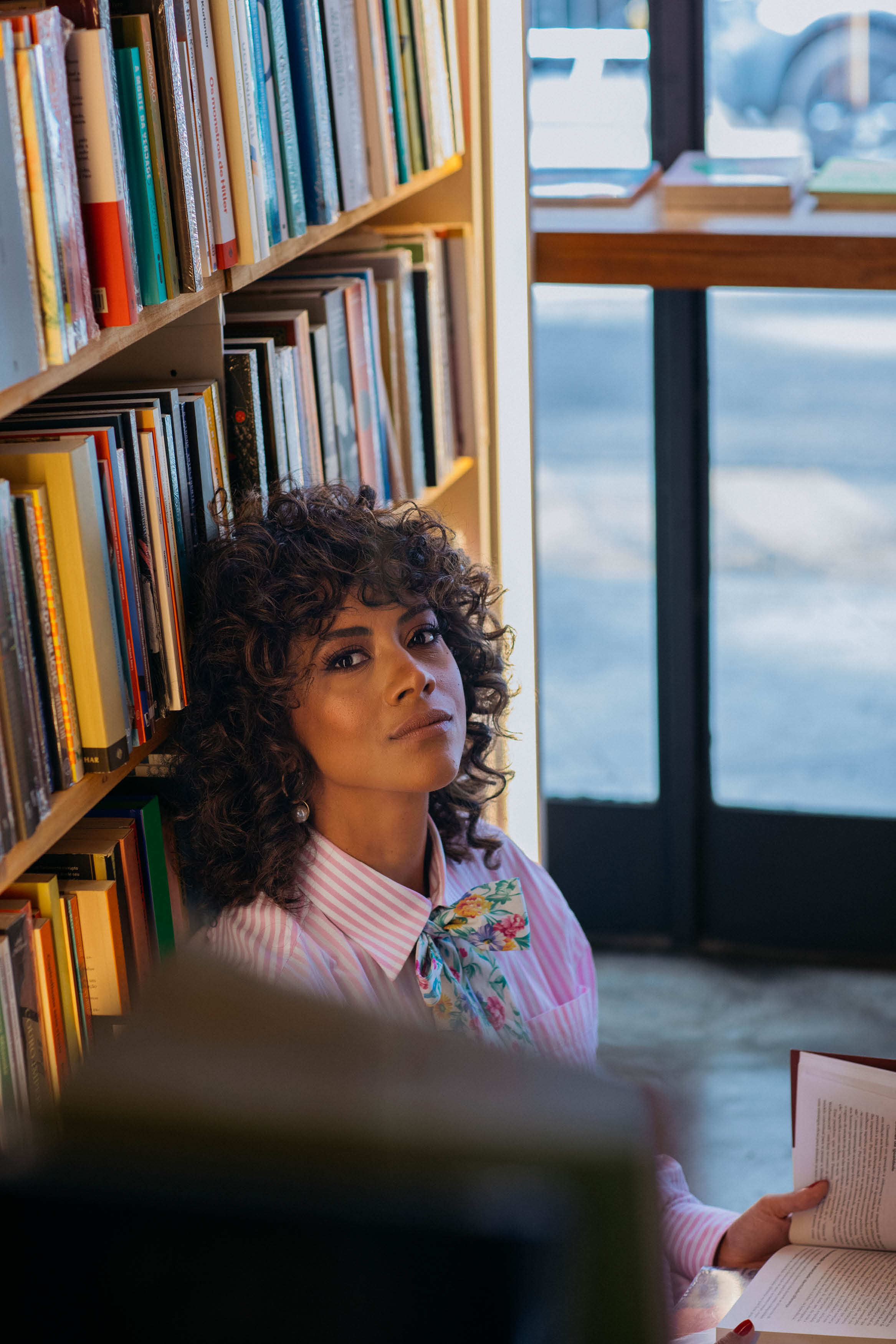Mulher sentada no ch&atilde;o de livraria encostada em estante de livros com um livro na m&atilde;o.