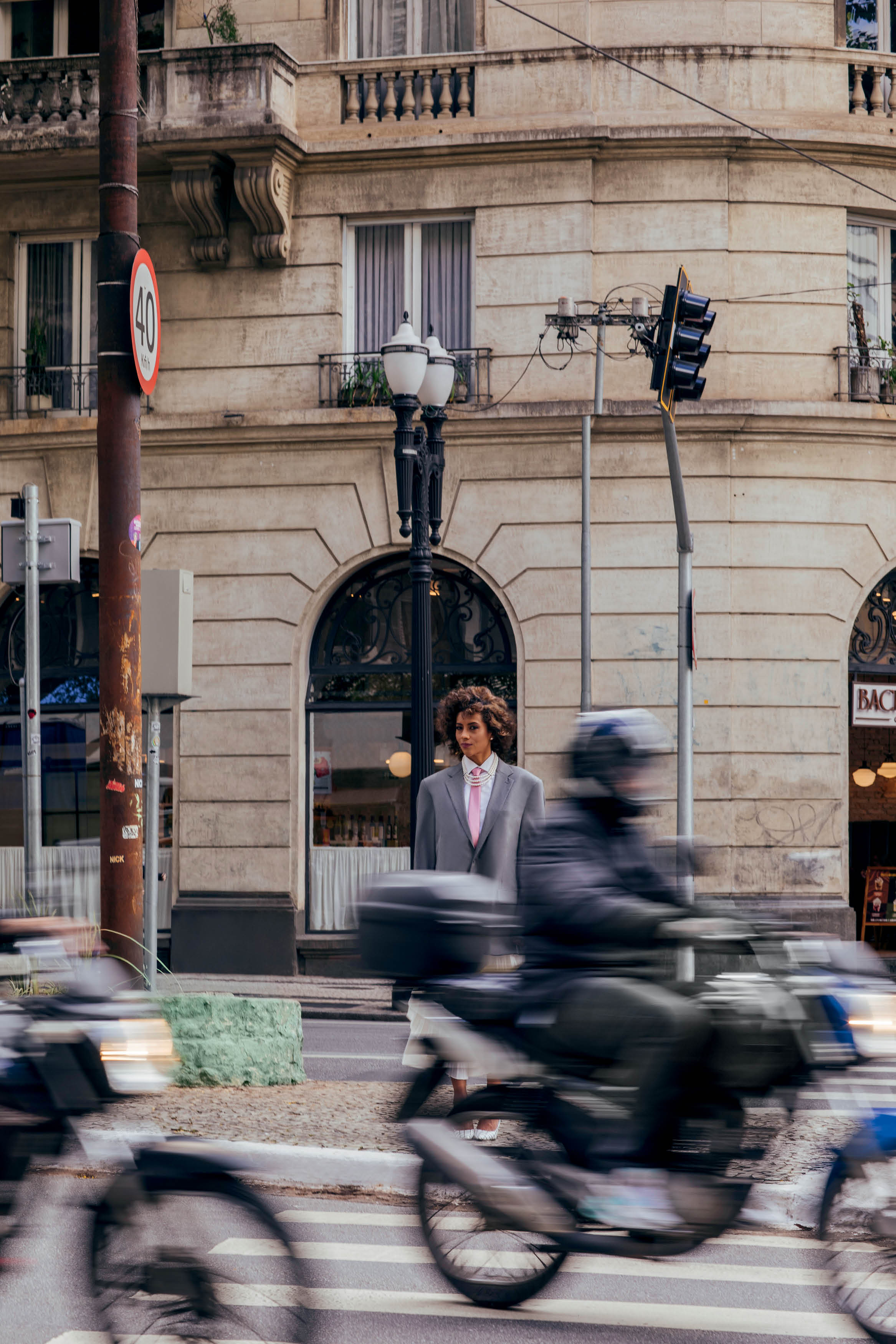 Mulher vestindo blazer cinza posando no meio da rua com motos em movimento.