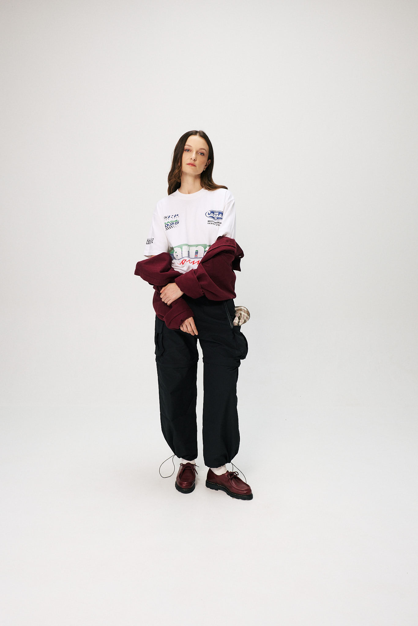 Modelo veste camiseta branca da collab Heineken 0.0 e Carnan, com estampa inspirada na F&oacute;rmula 1, cal&ccedil;a preta cargo e jaqueta vinho, em est&uacute;dio de fundo branco.