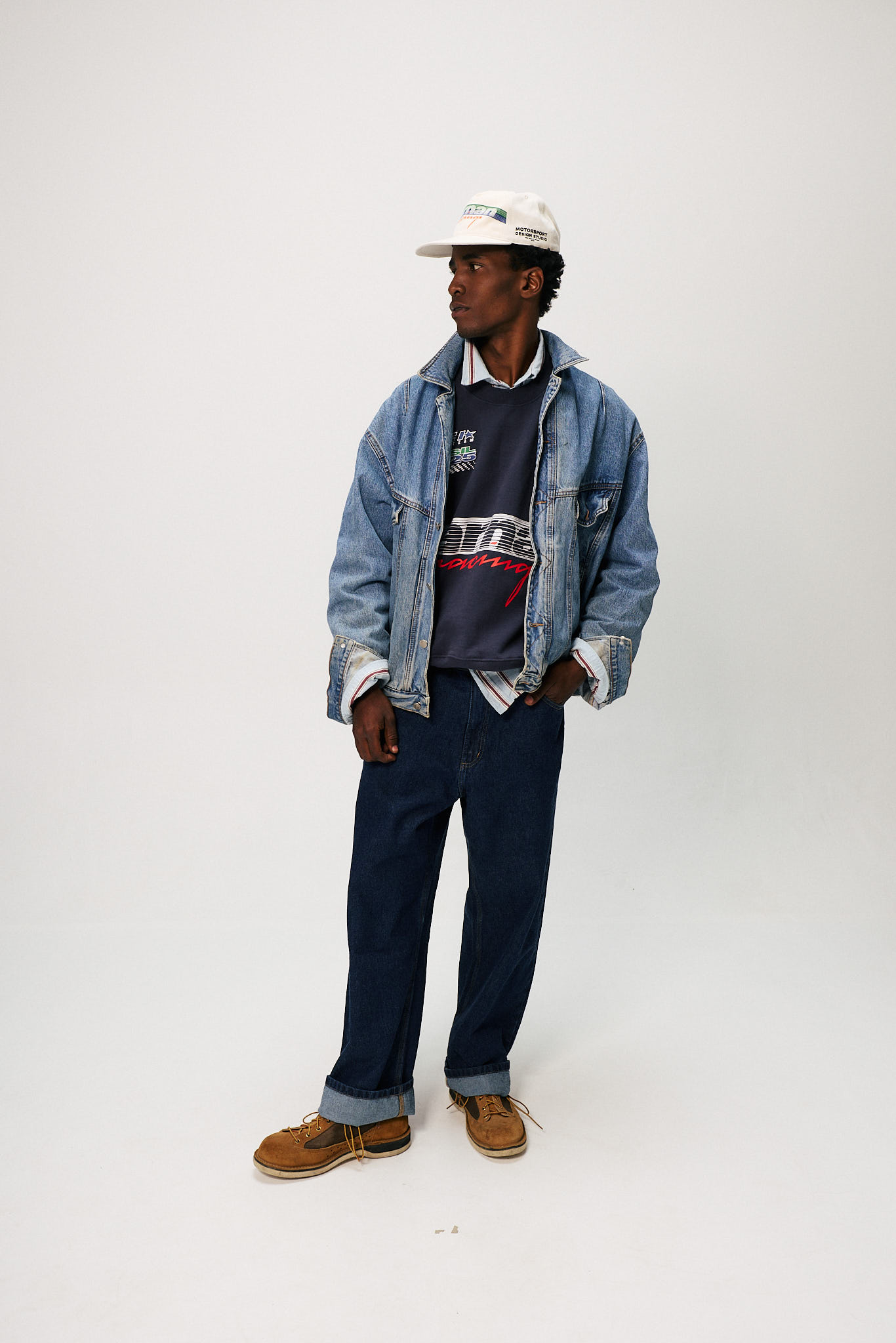 Modelo masculino veste look da collab Heineken 0.0 e Carnan composto por moletom azul com estampa racing, camisa listrada, jaqueta jeans oversized, cal&ccedil;a jeans escura dobrada na barra e bon&eacute; branco