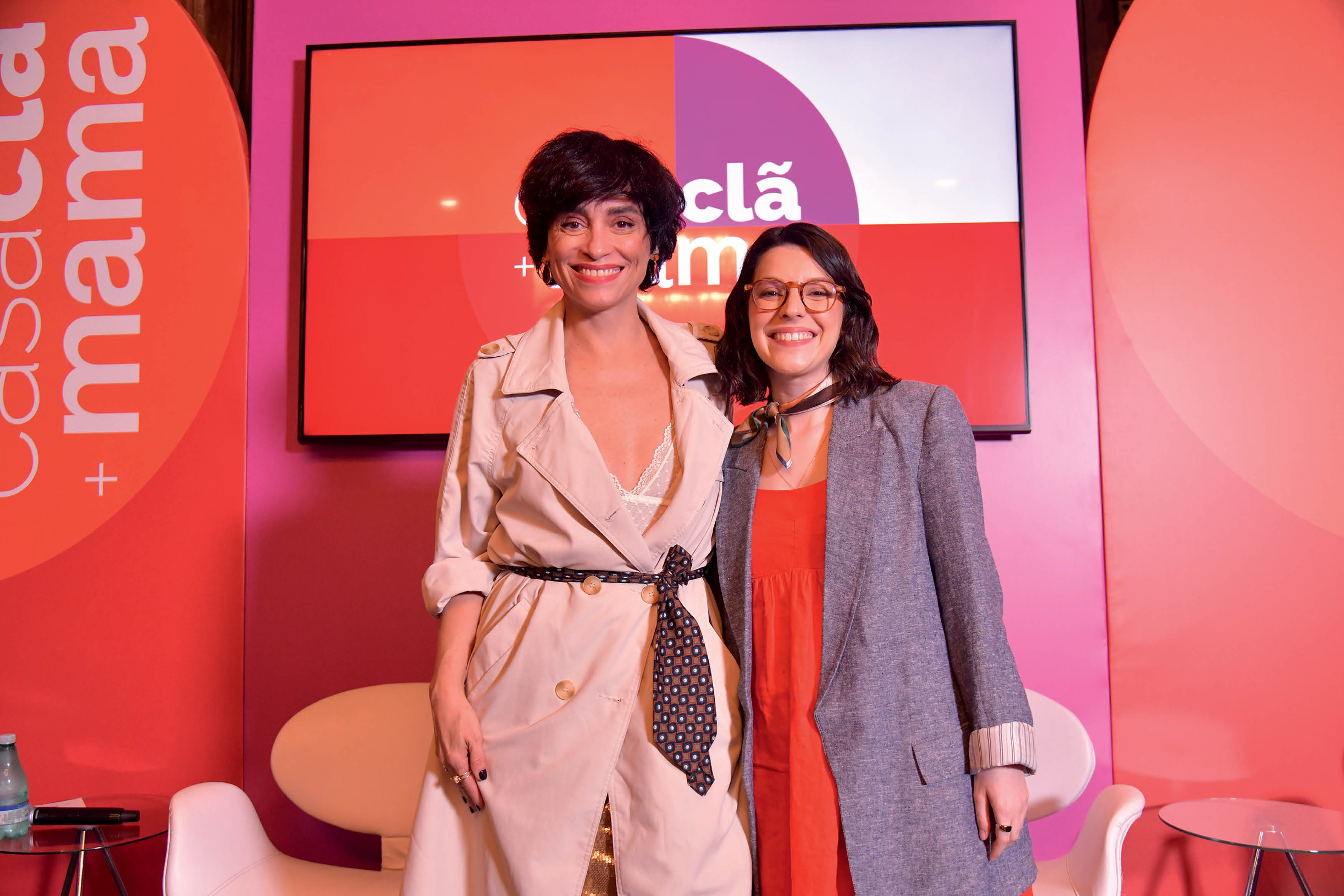 Duas mulheres posando para foto lado a lado &agrave; frente de palco com tema rosa.