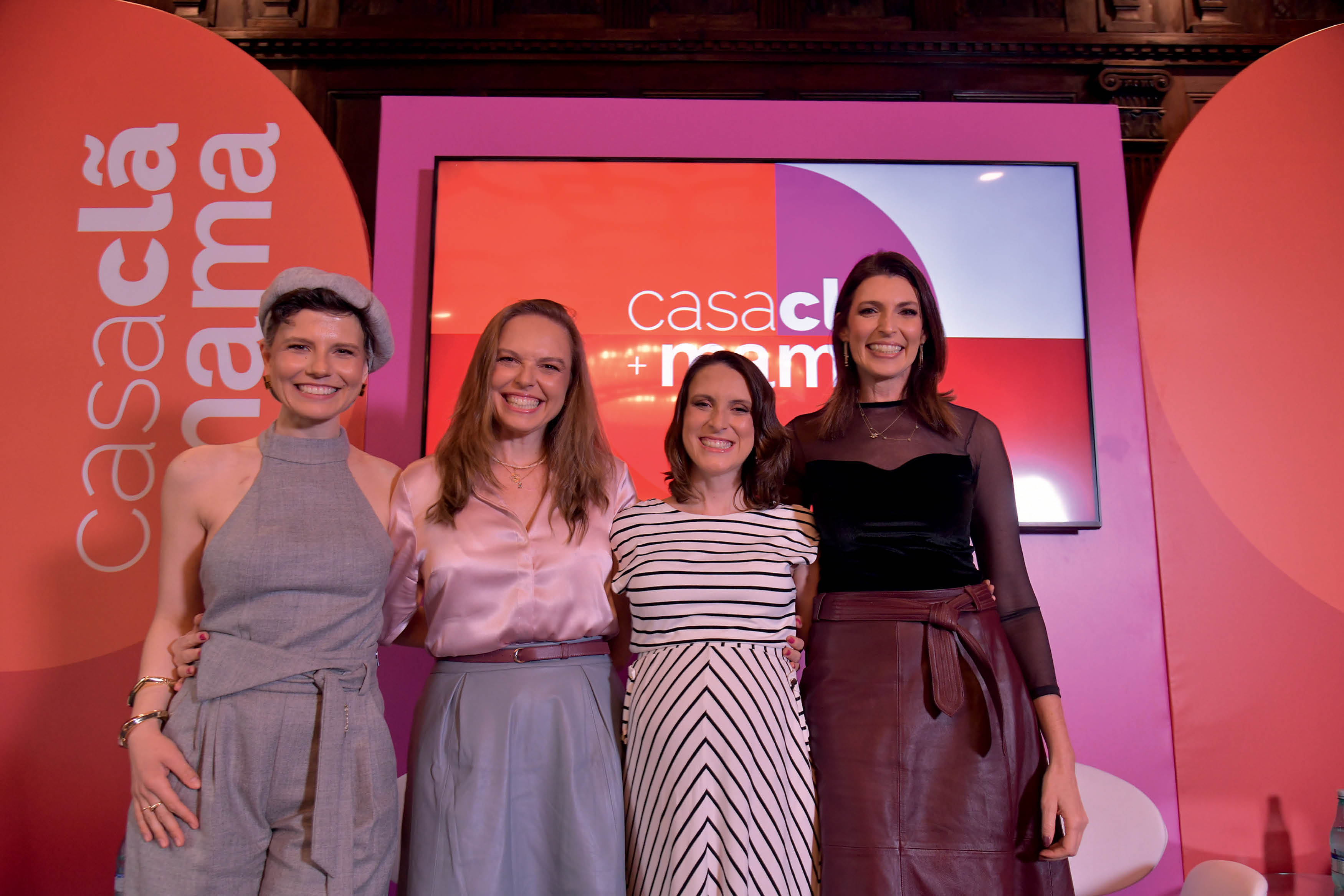 Quatro mulheres posando lado a lado em palco de evento com fundo rosa.