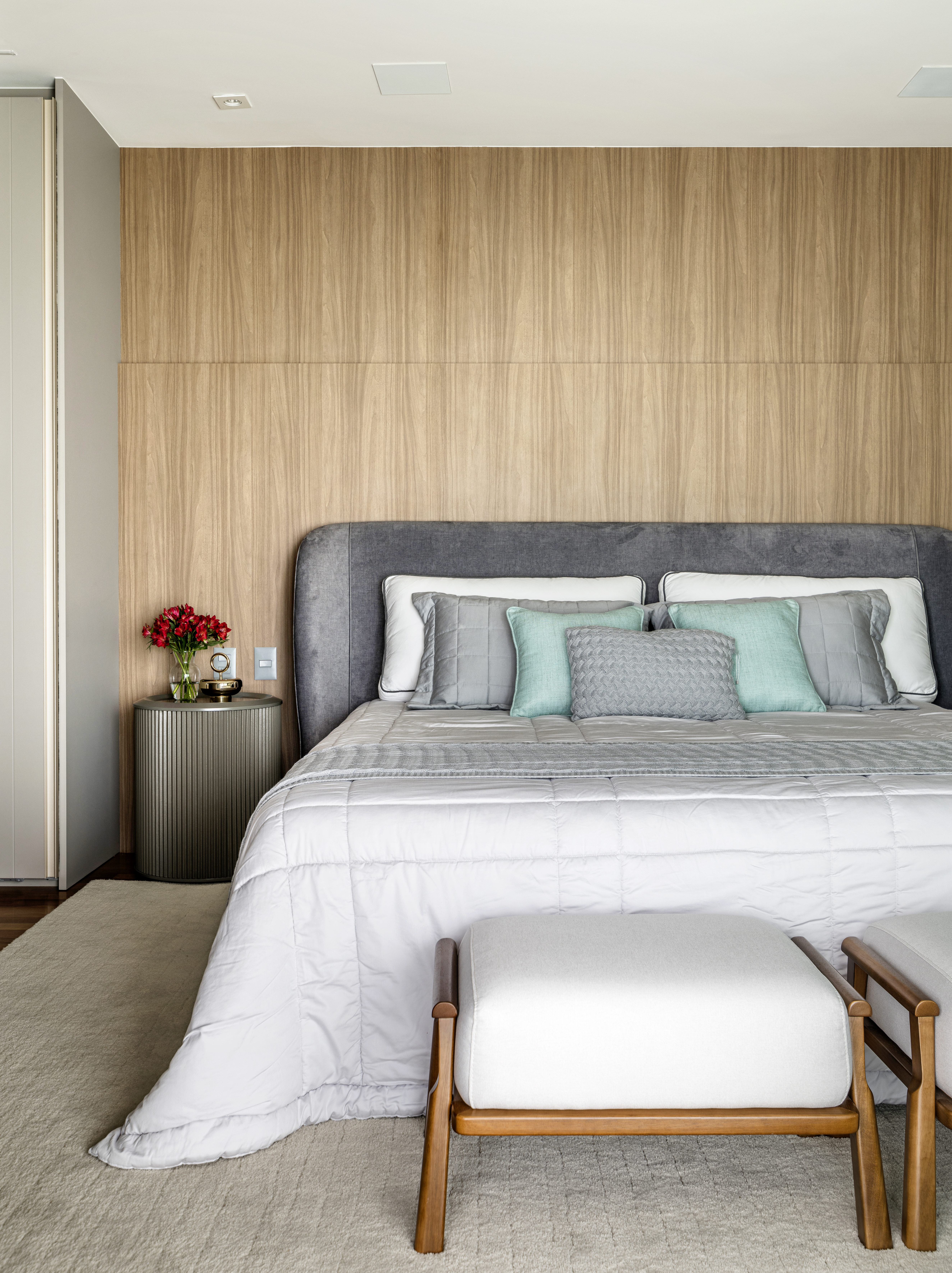 Quarto com parede de madeira, cama de casal com roupa de cama branca, azul e cinza, e brancos brancos em frente &agrave; cama.