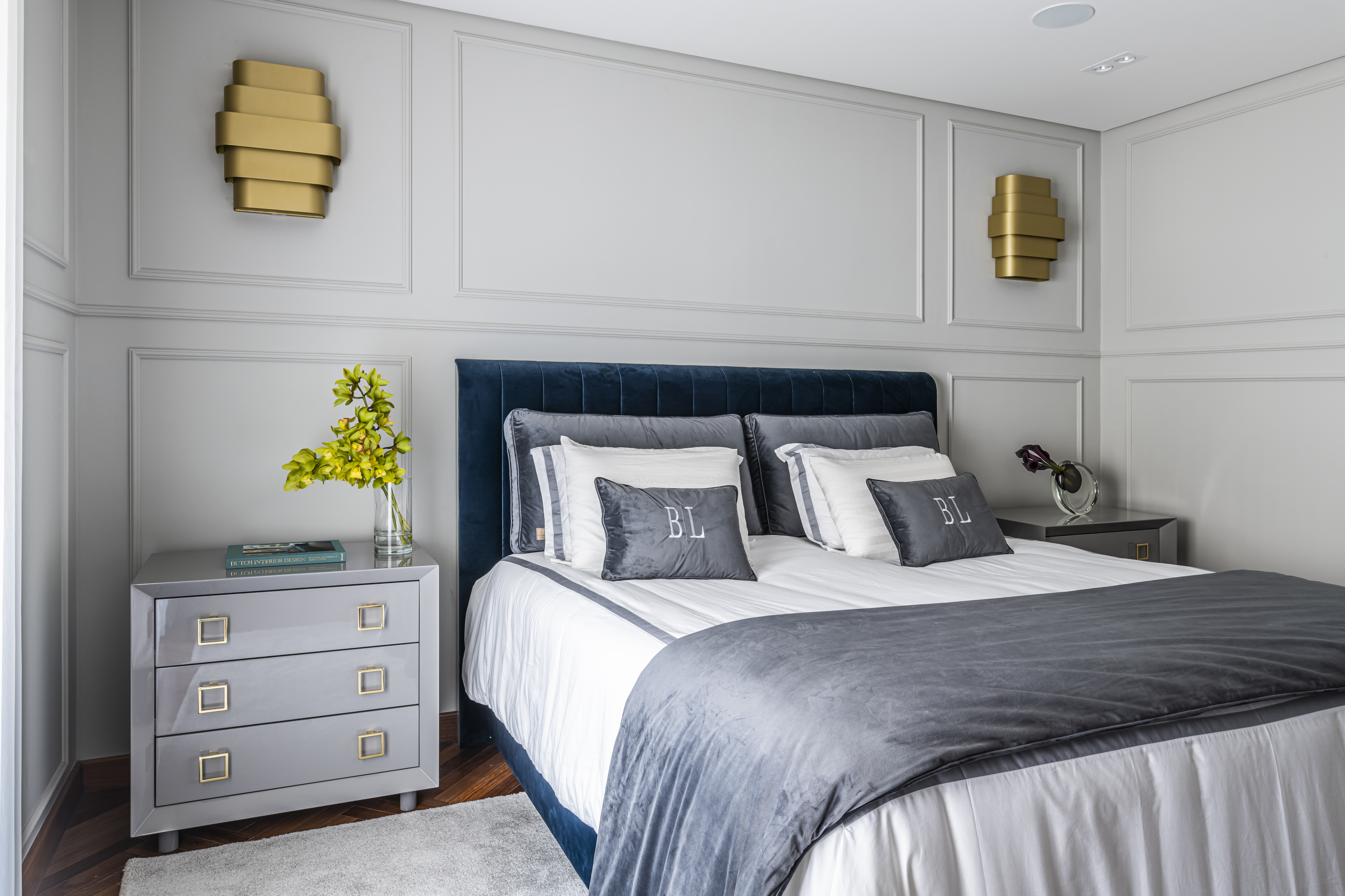 Quarto de casal com decora&ccedil;&atilde;o branca, lumin&aacute;rias douradas e roupa de cama azul escuro e cinza.