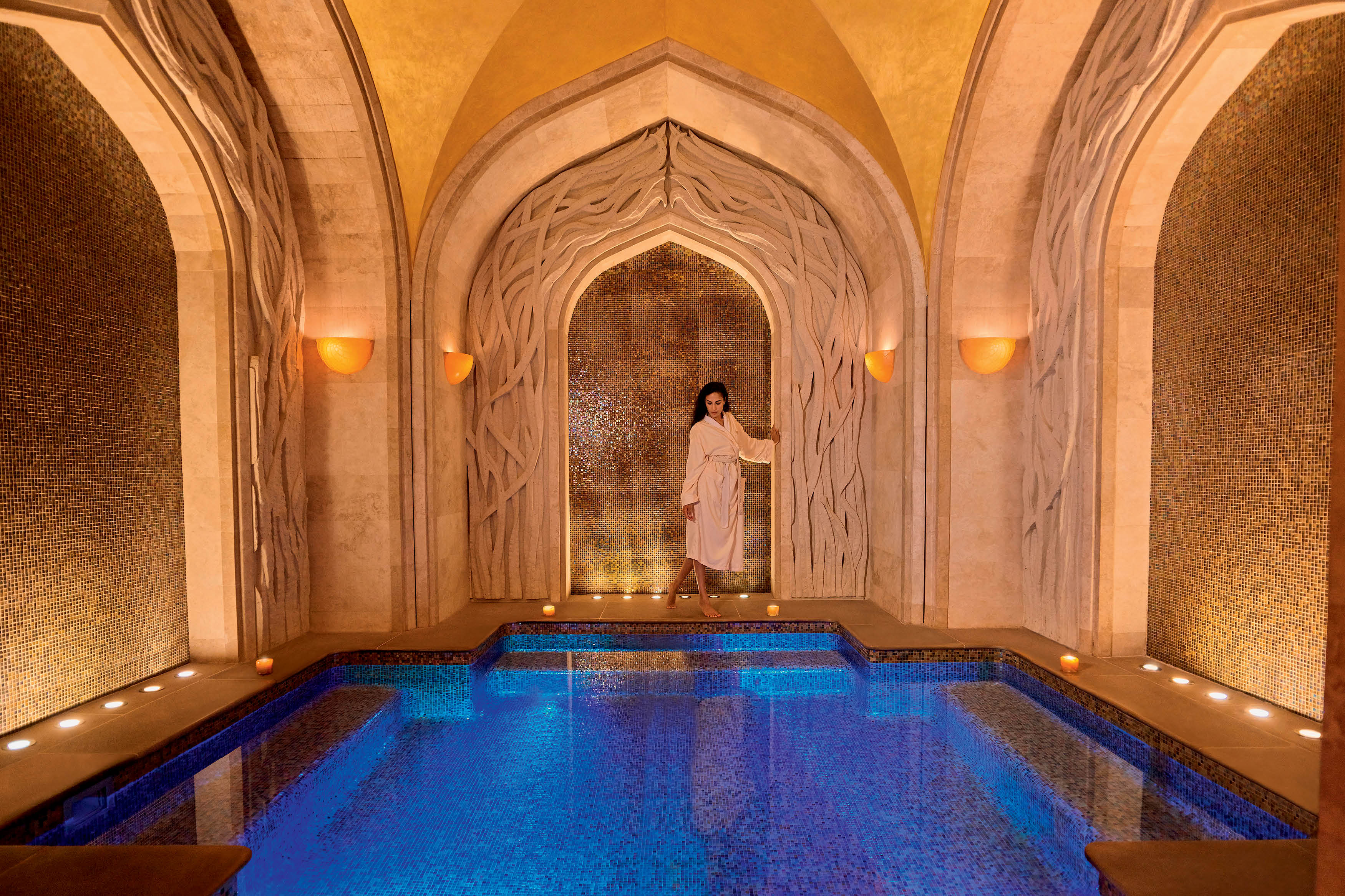 Spa luxuoso com arquitetura dos Emirados &Aacute;rabes com piscina azul e mulher em p&eacute; vestindo roup&atilde;o branco.