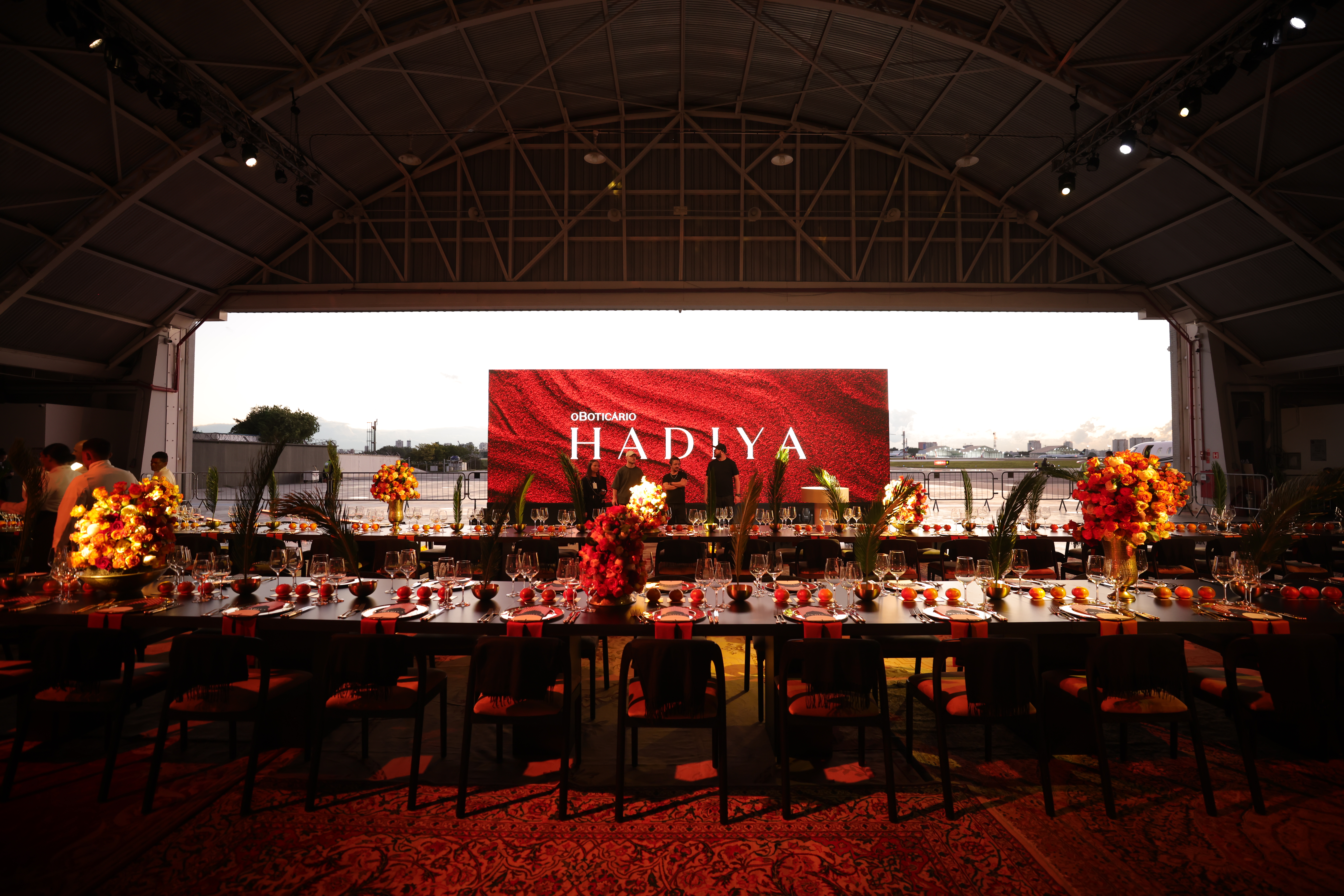 Ambiente do evento de lan&ccedil;amento de Hadiya em S&atilde;o Paulo, com mesas decoradas em tons quentes, arranjos florais e painel ao fundo com o nome do perfume. Pessoas circulam pelo local, incluindo convidados como Silvia Braz, o perfumista Adilson Rato e a VP de marketing Renata Gomide.