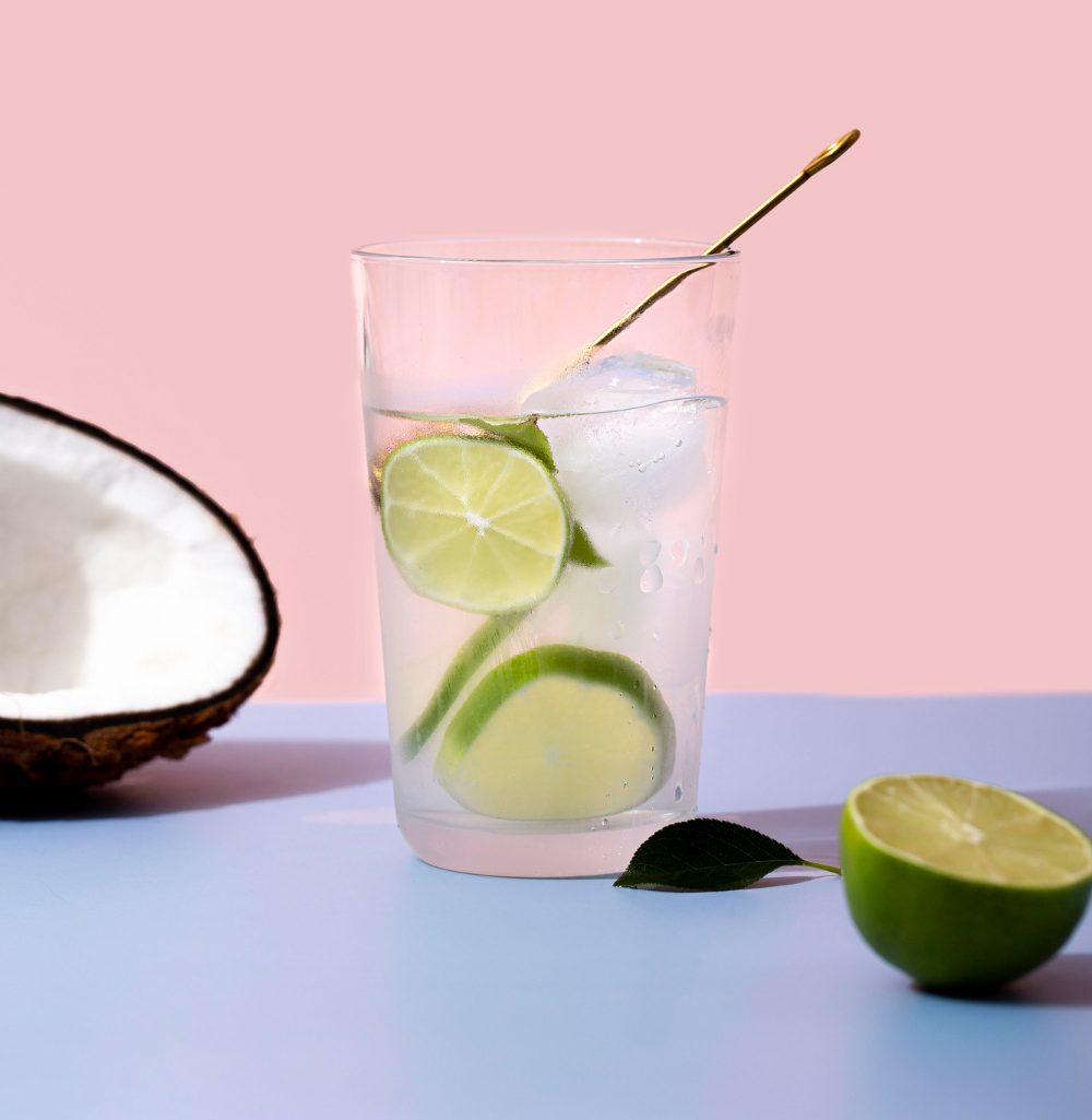 Copo de vidro com uma bebida clara (representando a &Aacute;gua de Coco) contendo rodelas de lim&atilde;o e gelo, sugerindo um shot de imunidade refrescante. Ao lado, metade de um coco e meio lim&atilde;o cortado confirmam os ingredientes. A bebida refere-se &agrave; receita de &Aacute;gua de Coco com Espirulina e Lim&atilde;o, que hidrata e fornece Vitamina C e ficocianina para fortalecer a imunidade.