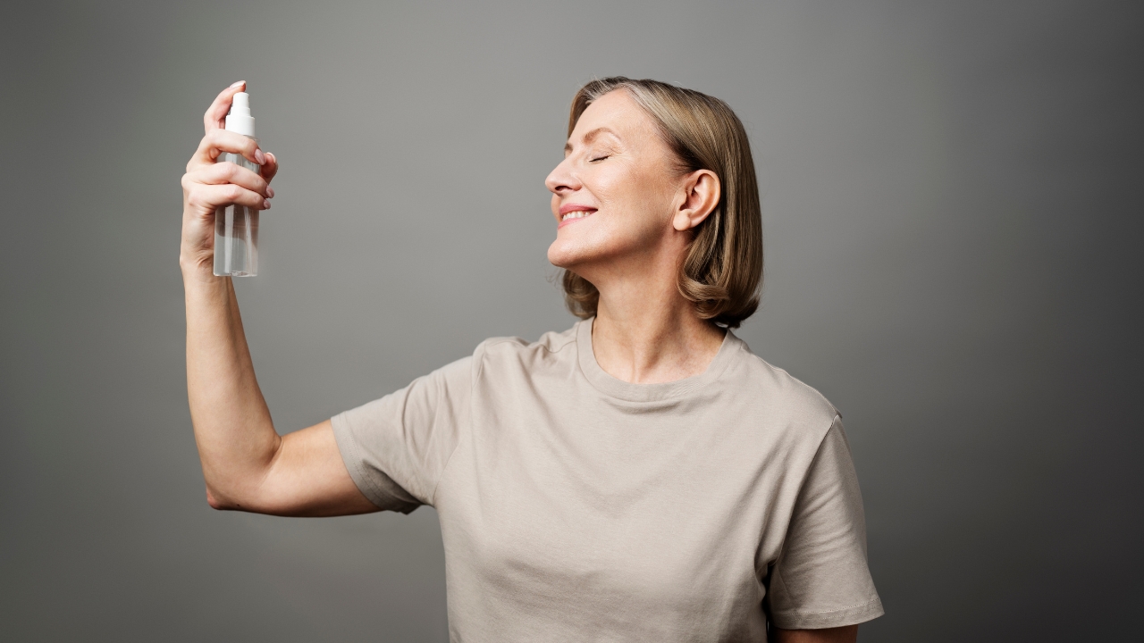 Mulher na faixa dos 50 anos com camisa cinza passando spray no rosto em fundo neutro de estúdio.