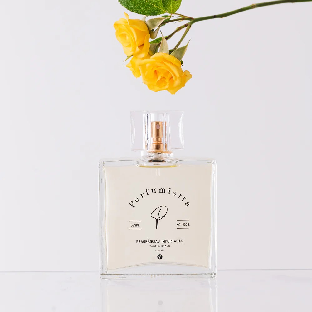 Foto de um perfume com a embalagem quadrada. H&aacute; tamb&eacute;m uma flor amarela no topo