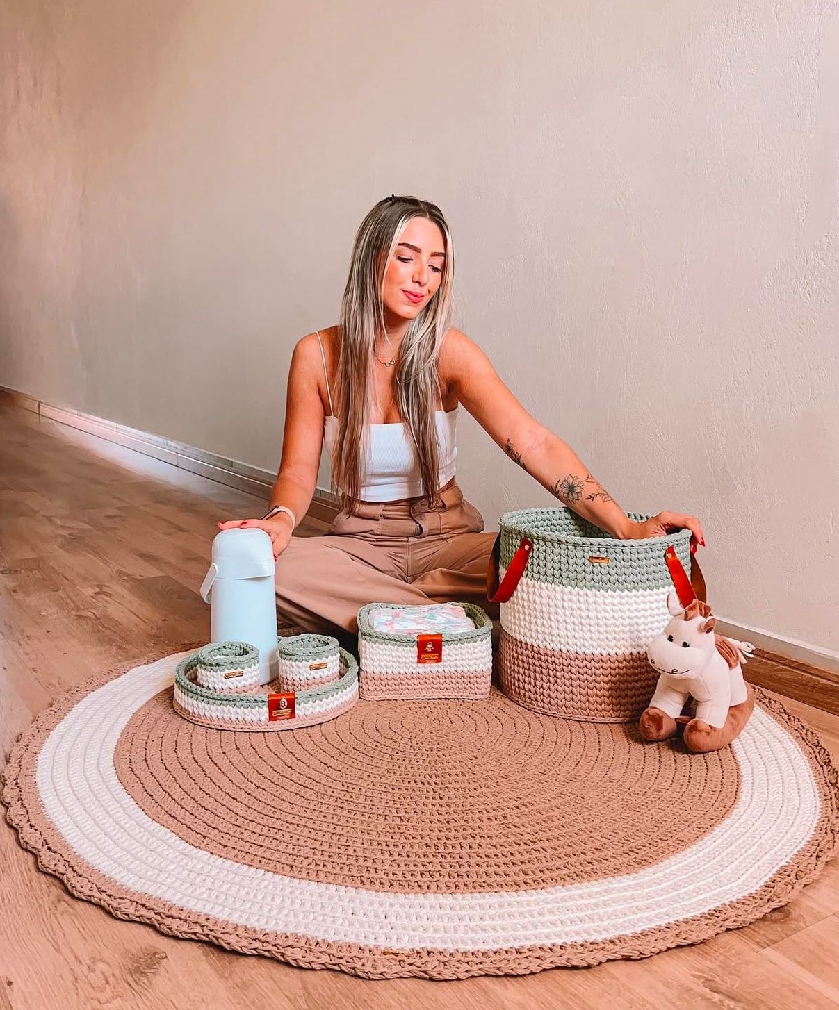 Foto de uma mulher loira sentada no ch&atilde;o. Ela toca um tapete infantil de croch&ecirc; marrom e branco. Tamb&eacute;m h&aacute; cestinhas de decora&ccedil;&atilde;o com o mesmo material