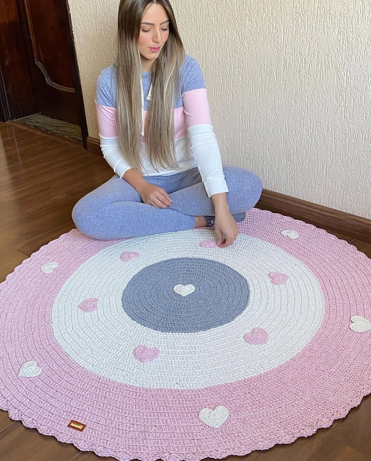 Foto de uma mulher loira sentada no ch&atilde;o. Ela toca um tapete infantil de croch&ecirc; rosa, branco e roxo, com cora&ccedil;&otilde;es