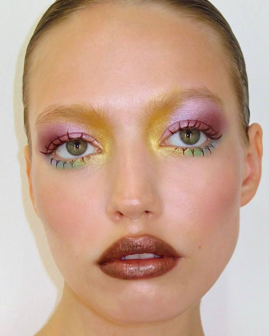 Foto de uma mulher loira usando uma maquiagem com sombra colorida. A sombra cont&eacute;m lil&aacute;s, roxo, amarelo e verde. Seu batom &eacute; marrom brilhante