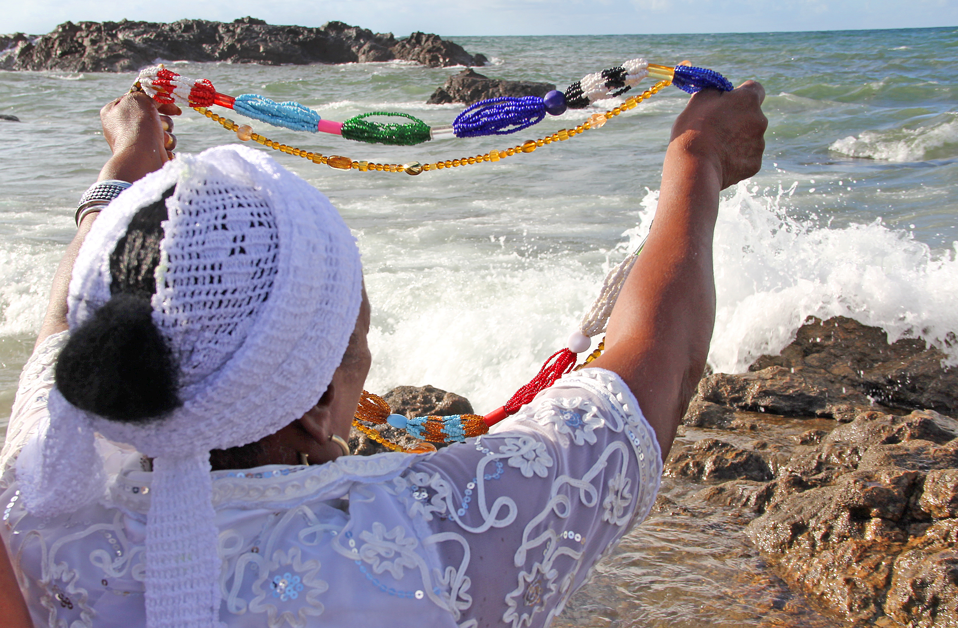 Pessoa de costas, vestida de branco e segurando um colar de contas coloridas em direção ao mar durante um ritual afro-religioso. A cena representa reverência aos Orixás e a busca por proteção e equilíbrio espiritual para 2026.