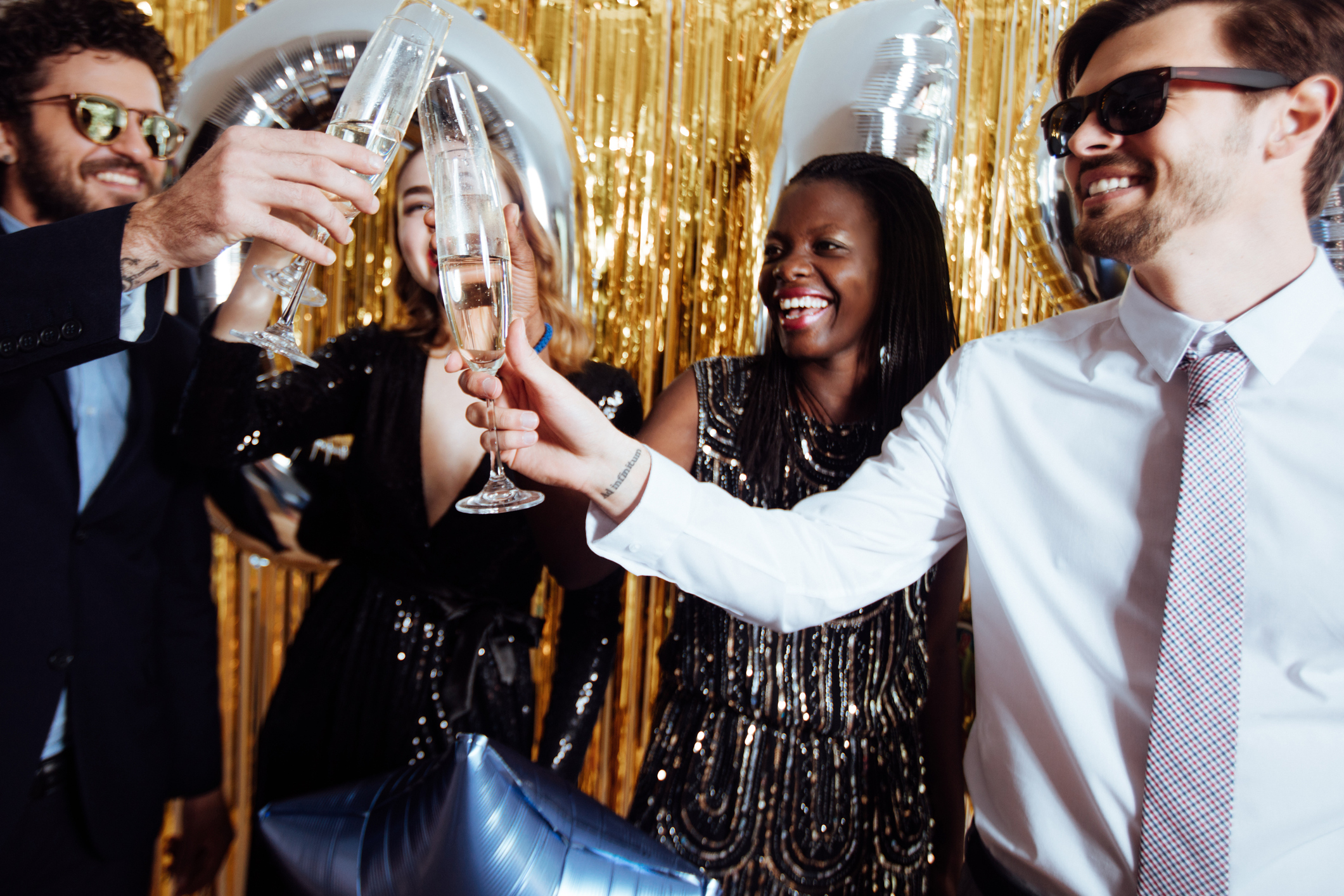 Grupo de amigos brindando em uma festa de Réveillon, com roupas elegantes em preto e dourado diante de um fundo festivo dourado, sorrindo e levantando taças de espumante.