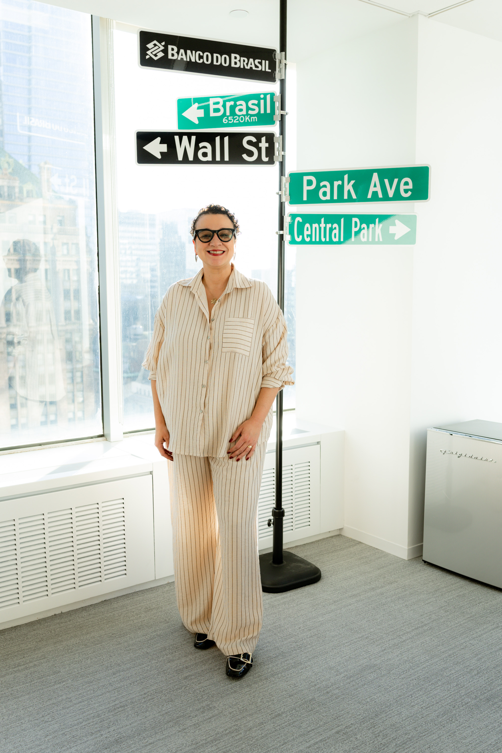 Foto de corpo inteiro de Tarciana Medeiros. Ela &eacute; morena, tem cabelo curto e cacheado, veste um conjunto de camisa e cal&ccedil;a de alfaiataria listrados e usa um &oacute;culos de grau