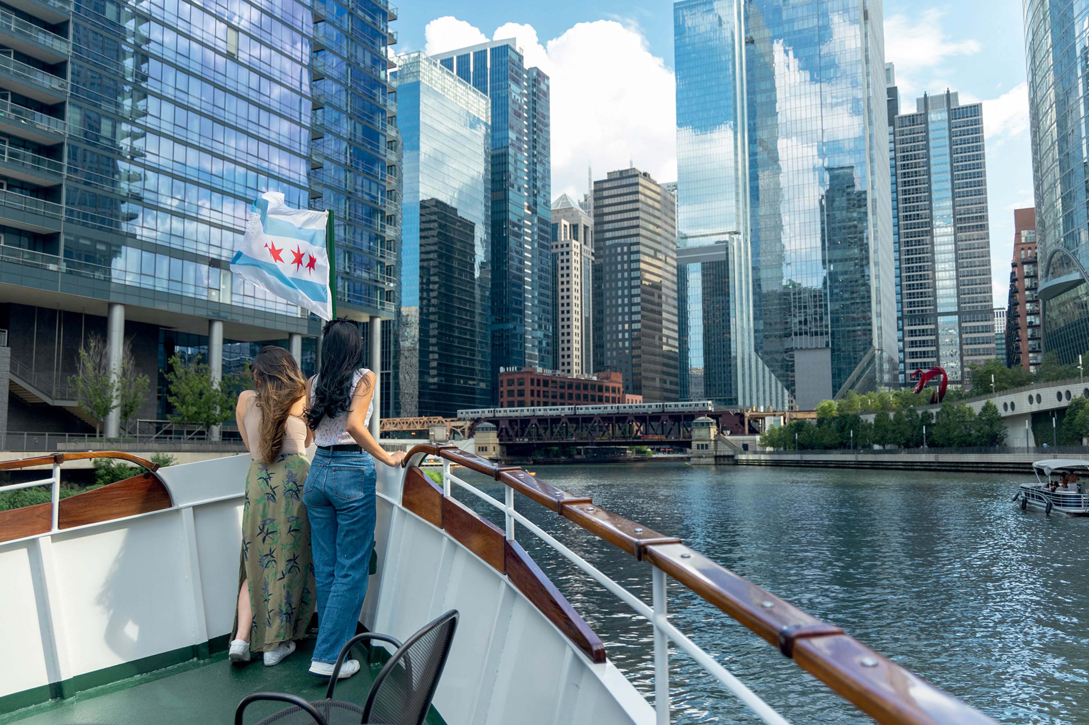 Turistas em cruzeiro no Rio Chicago observam o skyline de arranha-c&eacute;us; bandeira de Chicago tremula no barco em dia ensolarado.