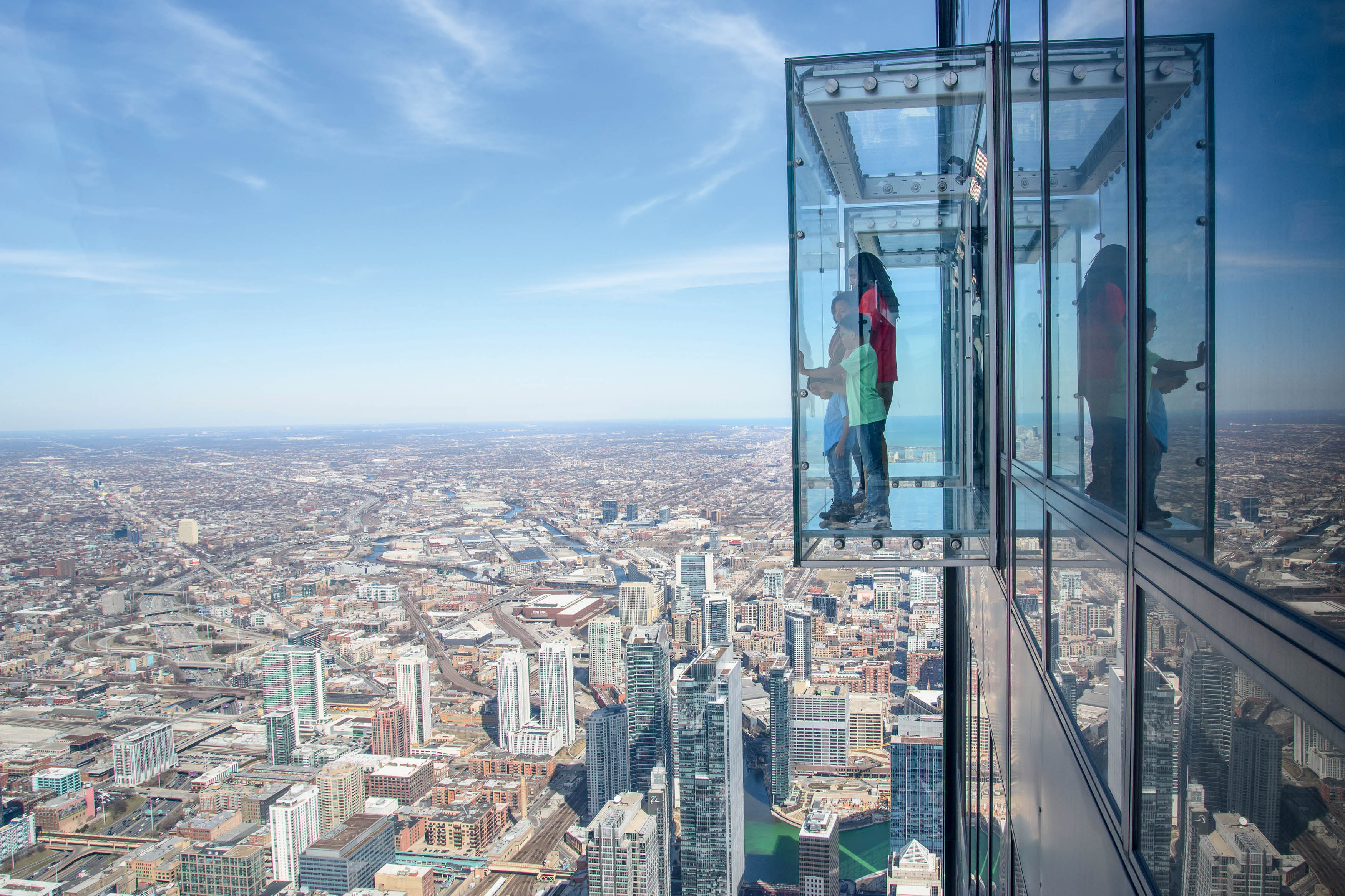 Visitantes em plataforma de vidro suspensa em mirante de Chicago, com vista vertiginosa da cidade e dos arranha-c&eacute;us em um dia de c&eacute;u azul