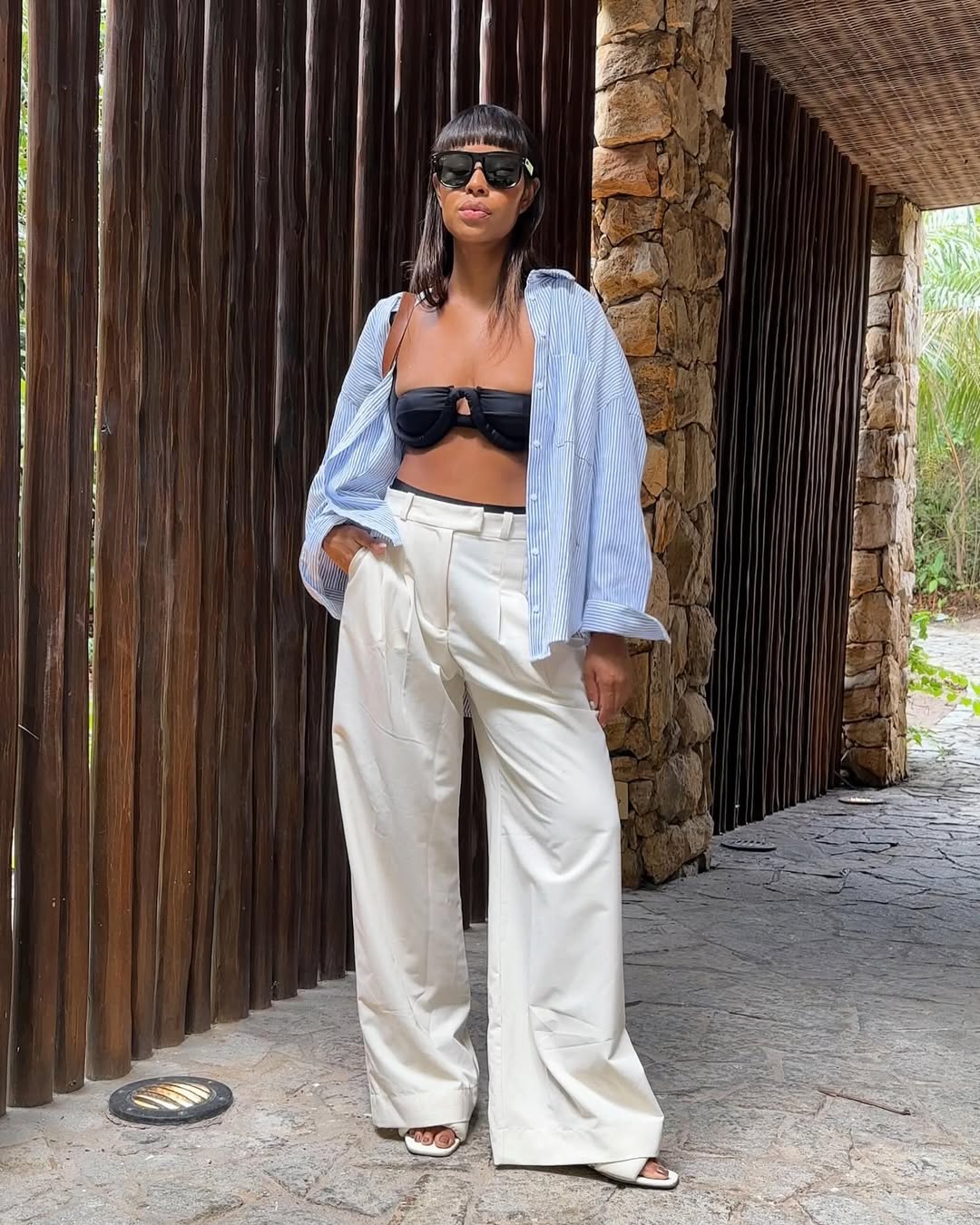 Foto de uma mulher negra usando um look composto por biquini preto, cal&ccedil;a branca de alfaiataria e camisa azul como terceira pe&ccedil;a